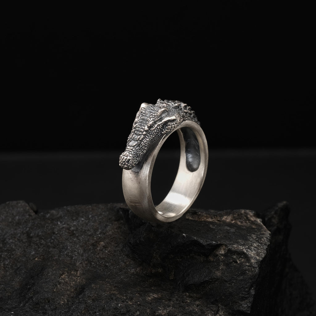 A silver ring with a crocodile head design is displayed on a rock against a black background.