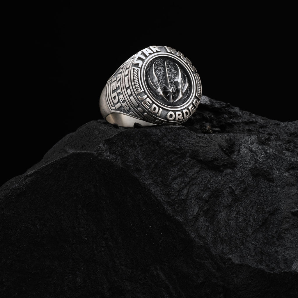 A silver ring with a Star Wars logo is placed on a black rock.