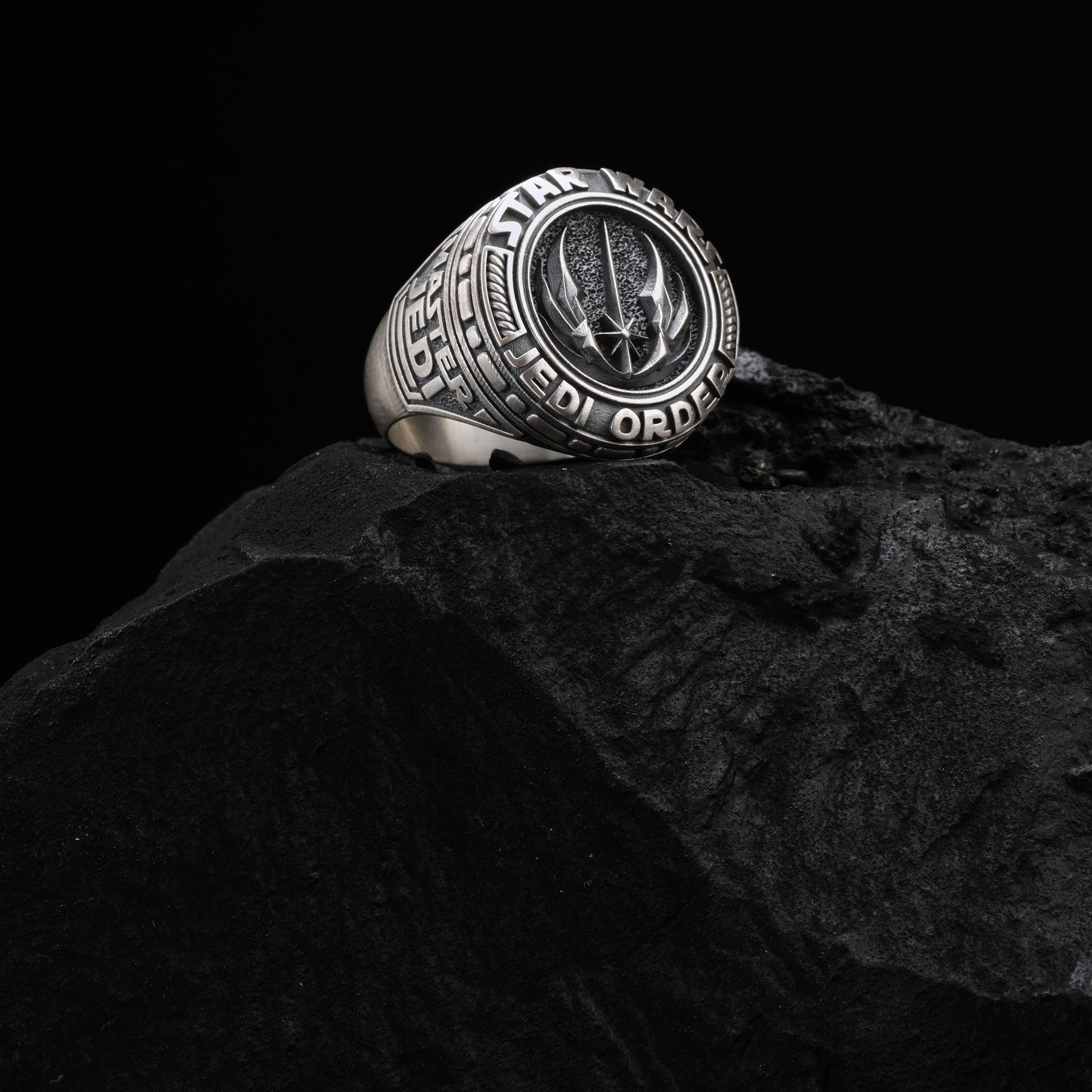 A silver ring with a Star Wars logo is placed on a black rock.