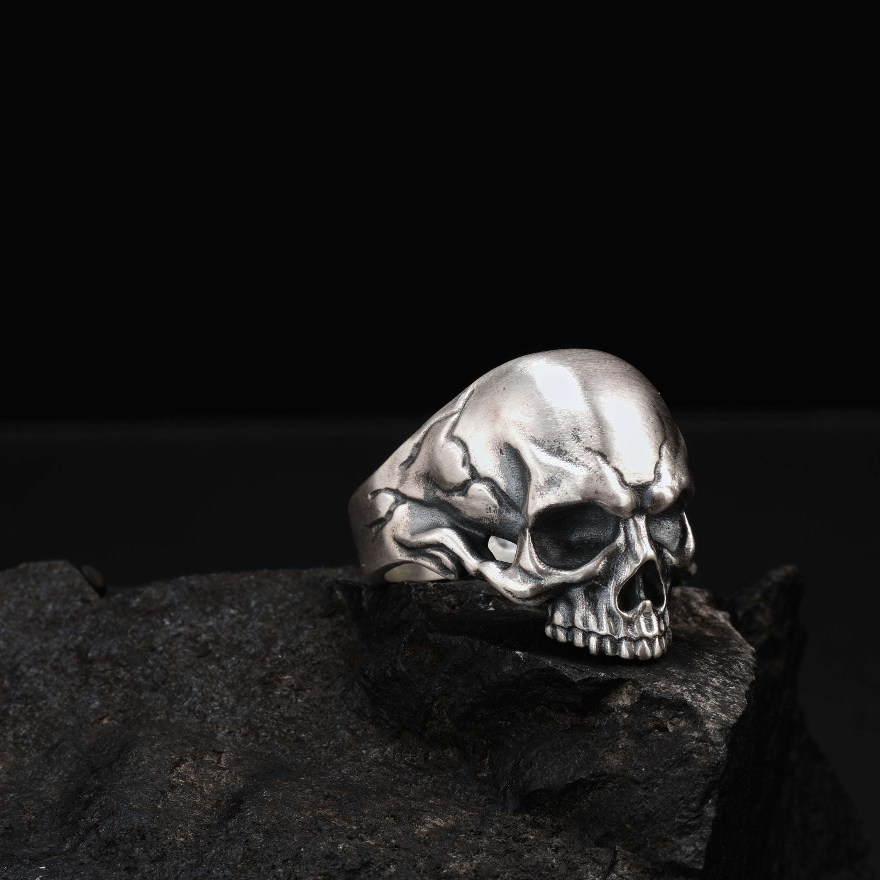 A silver skull ring is resting on a dark, rocky surface against a black background.