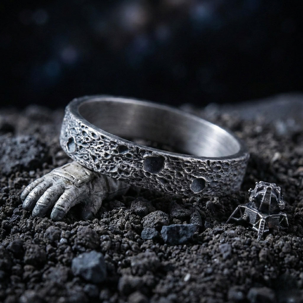 A silver ring with a textured surface sits on a bed of small rocks, with a small metal object resembling a spacecraft or lunar module nearby.