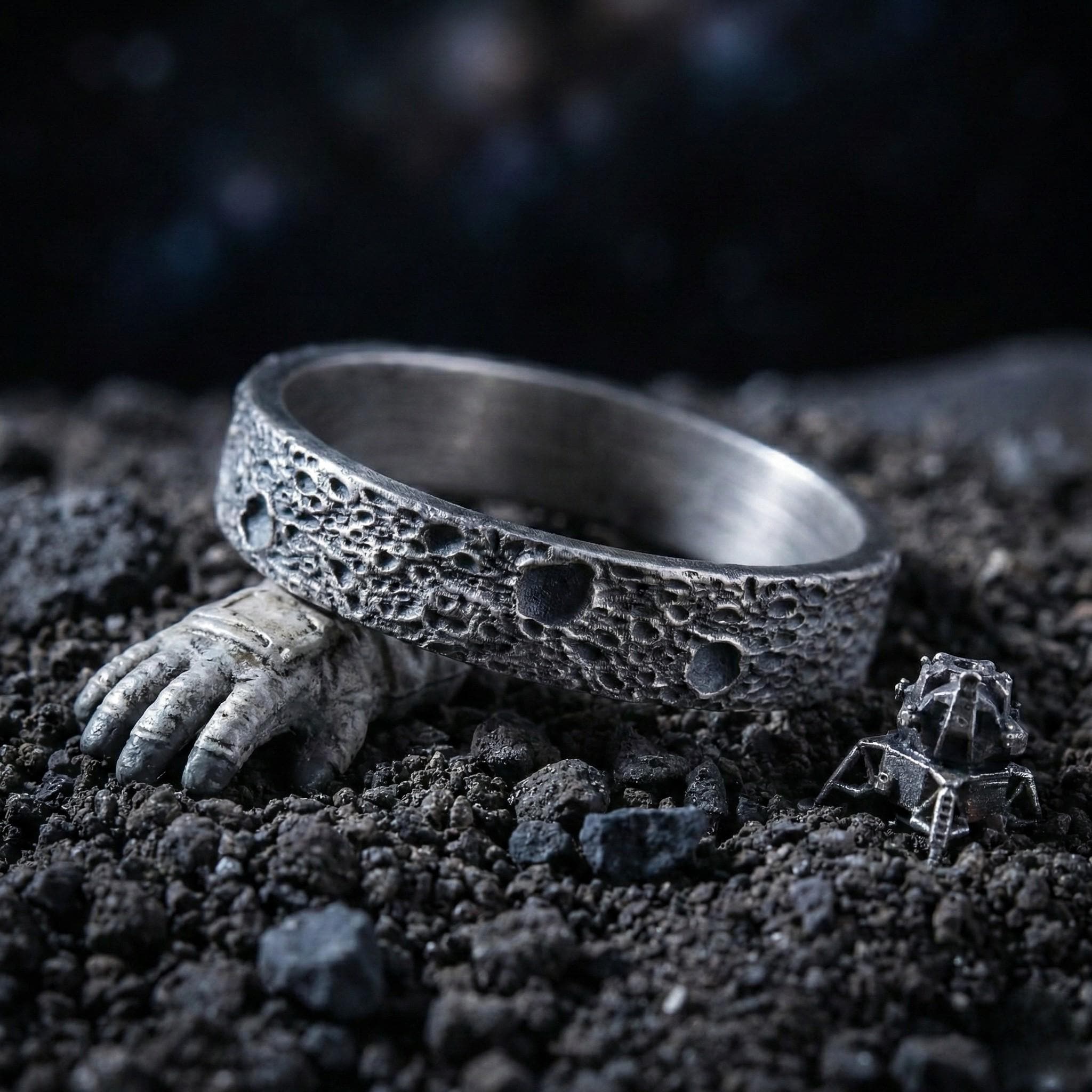 A silver ring with a textured surface sits on a bed of small rocks, with a small metal object resembling a spacecraft or lunar module nearby.