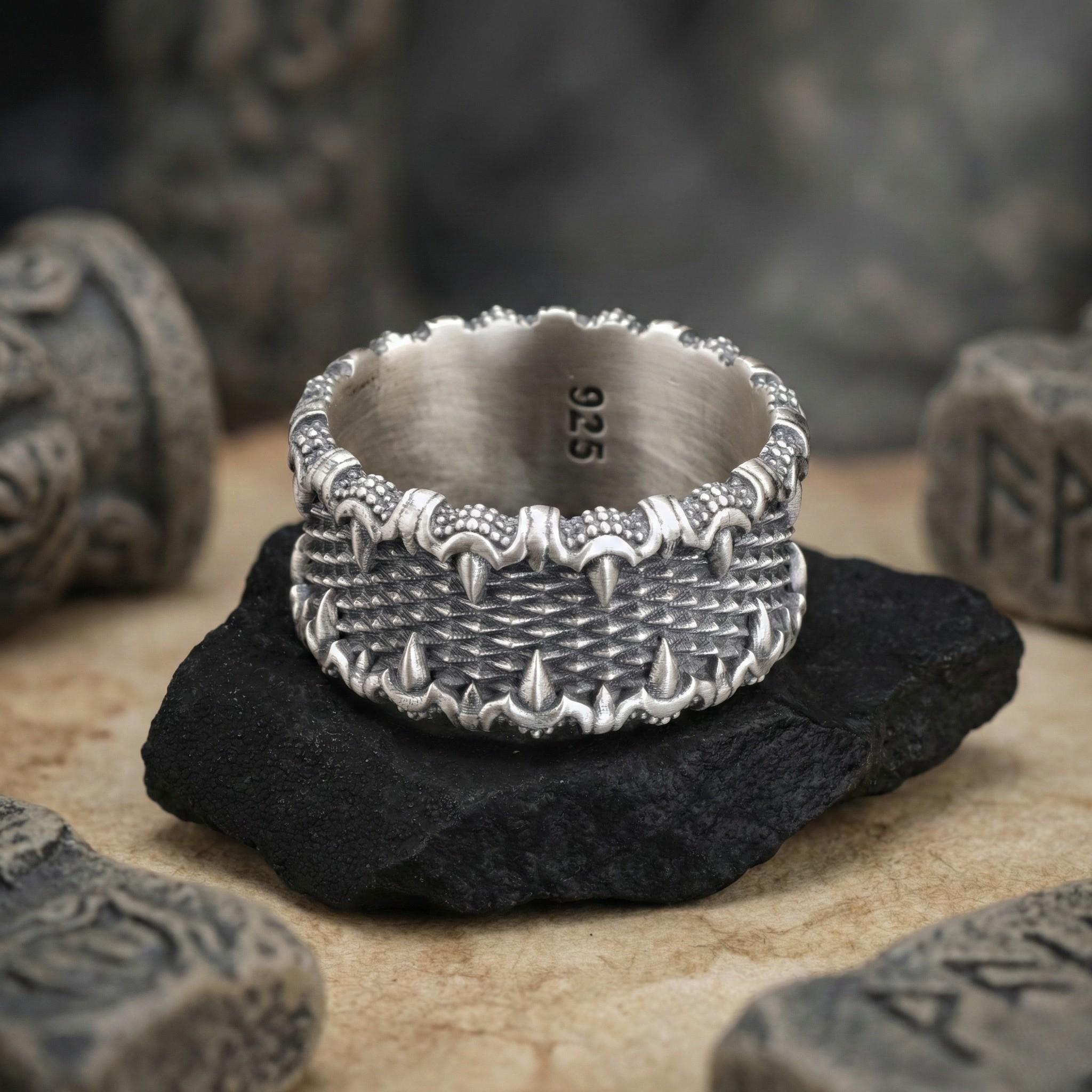 A silver ring with intricate designs sits on a black rock, surrounded by other objects with text on them.