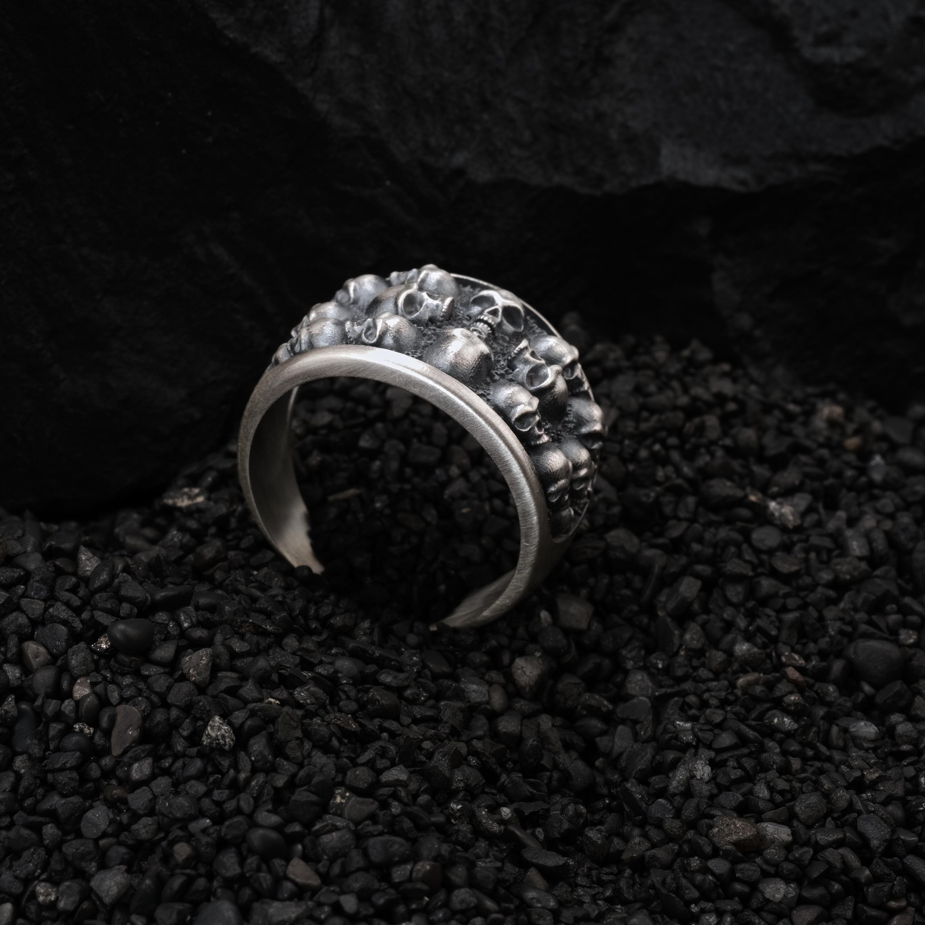 A silver ring with a skull design is resting on a bed of small black pebbles or stones.