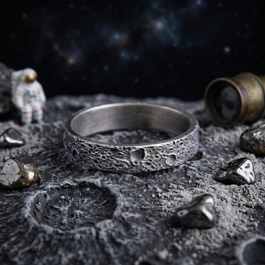 a ring with a moon-like texture, placed on a rocky surface with small rocks and a figurine of an astronaut in the background.