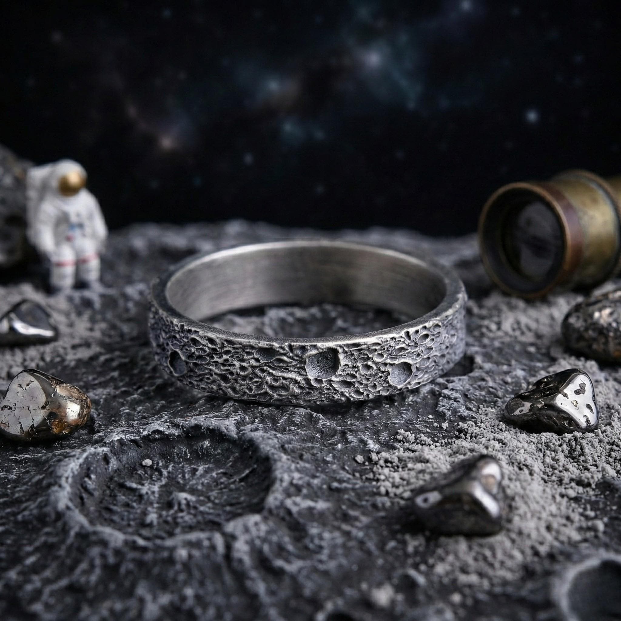 a ring with a moon-like texture, placed on a rocky surface with small rocks and a figurine of an astronaut in the background.