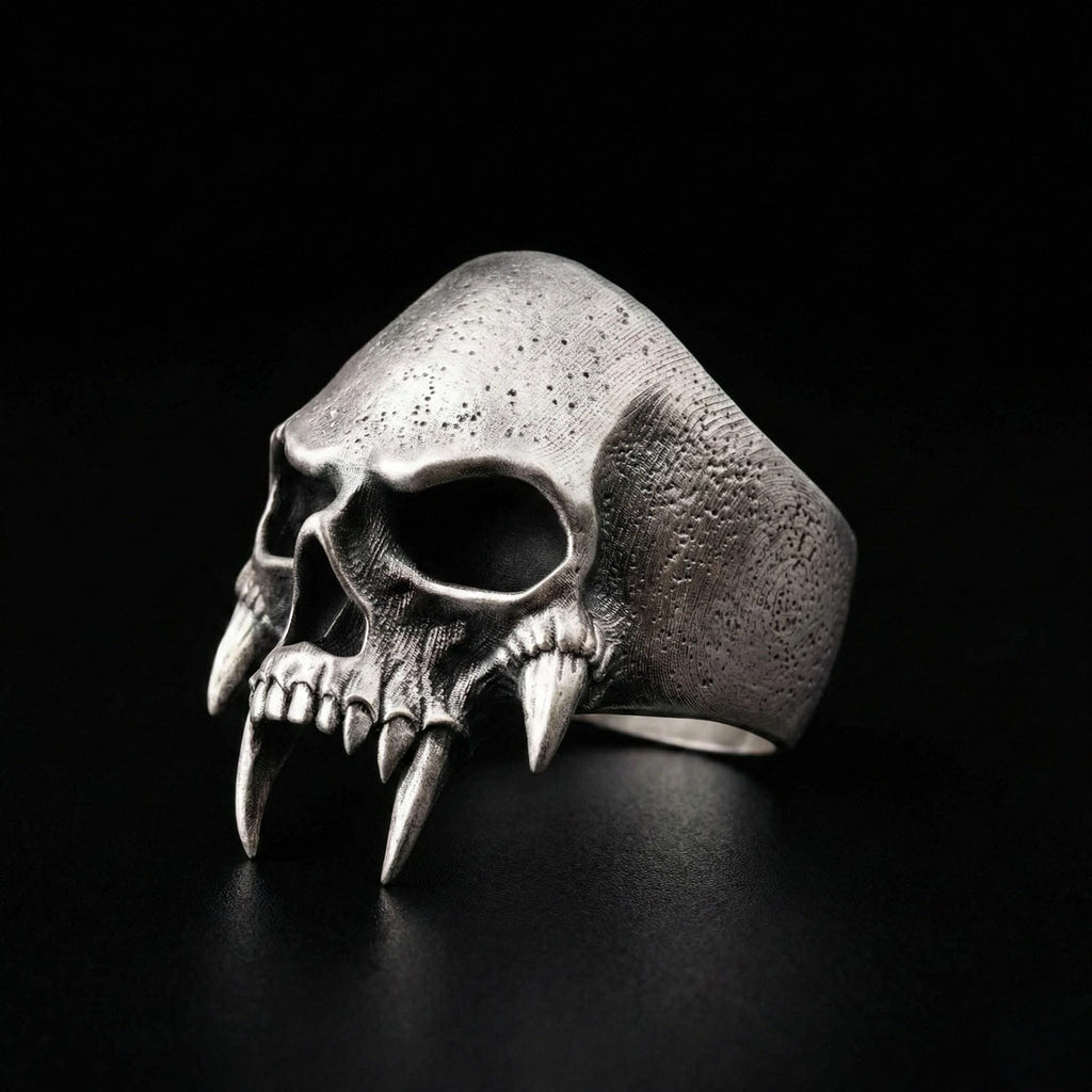 A silver ring with a skull design, featuring sharp teeth and a menacing expression, is shown against a black background.