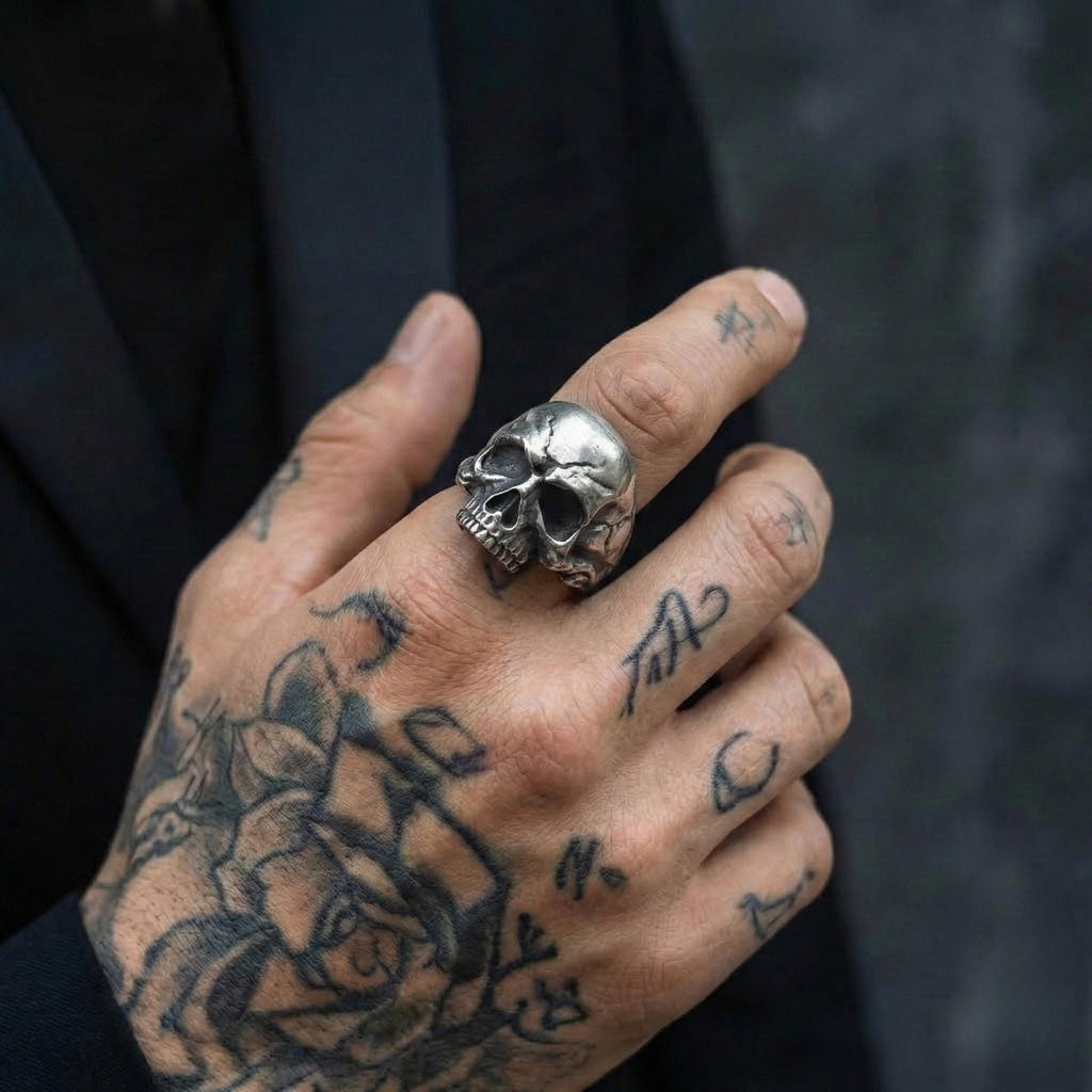 A close-up of a person's hand with a large skull ring on one finger and various tattoos on the other fingers.