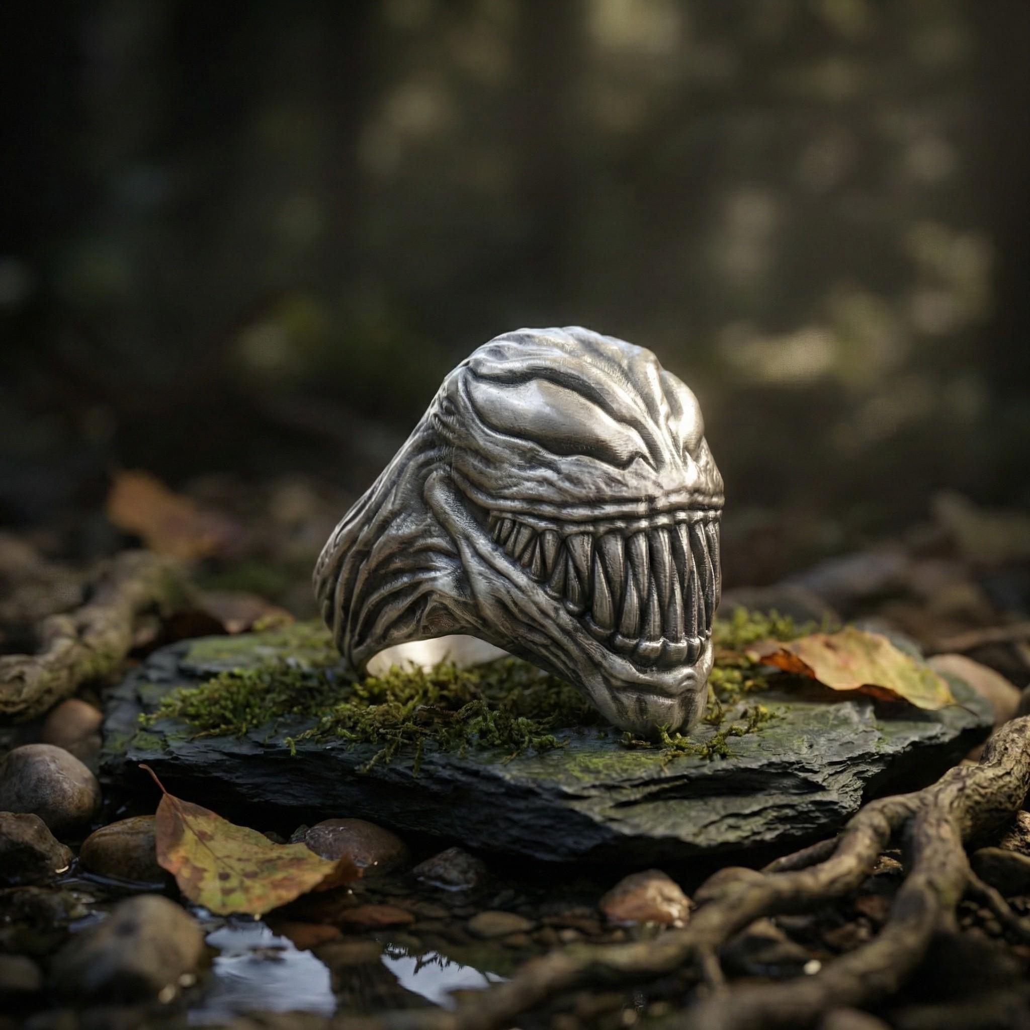 A silver ring with a large, menacing-looking creature face sits on a mossy rock in a forest setting.