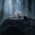 A silver ring with an intricate design sits on a black rock in a dark, wooded area.