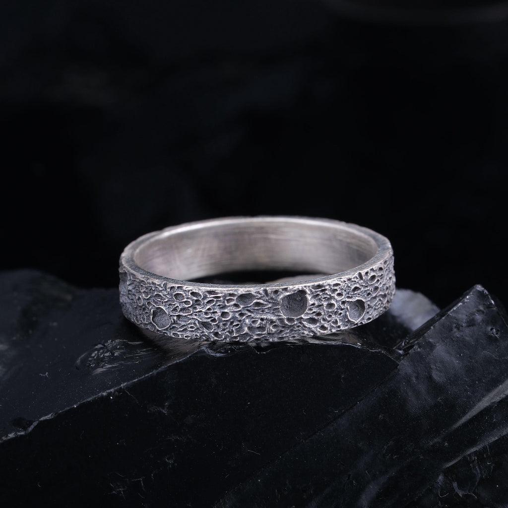 A silver ring with a textured surface sits on a black surface against a dark background.