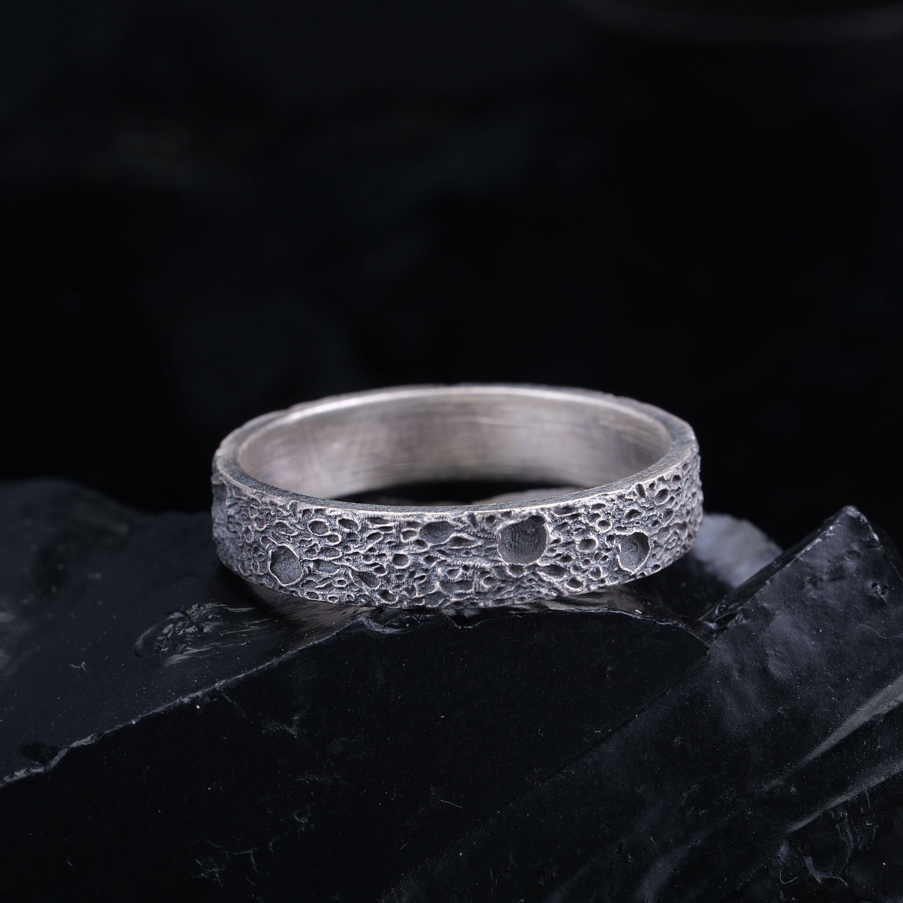A silver ring with a textured surface sits on a black surface against a dark background.