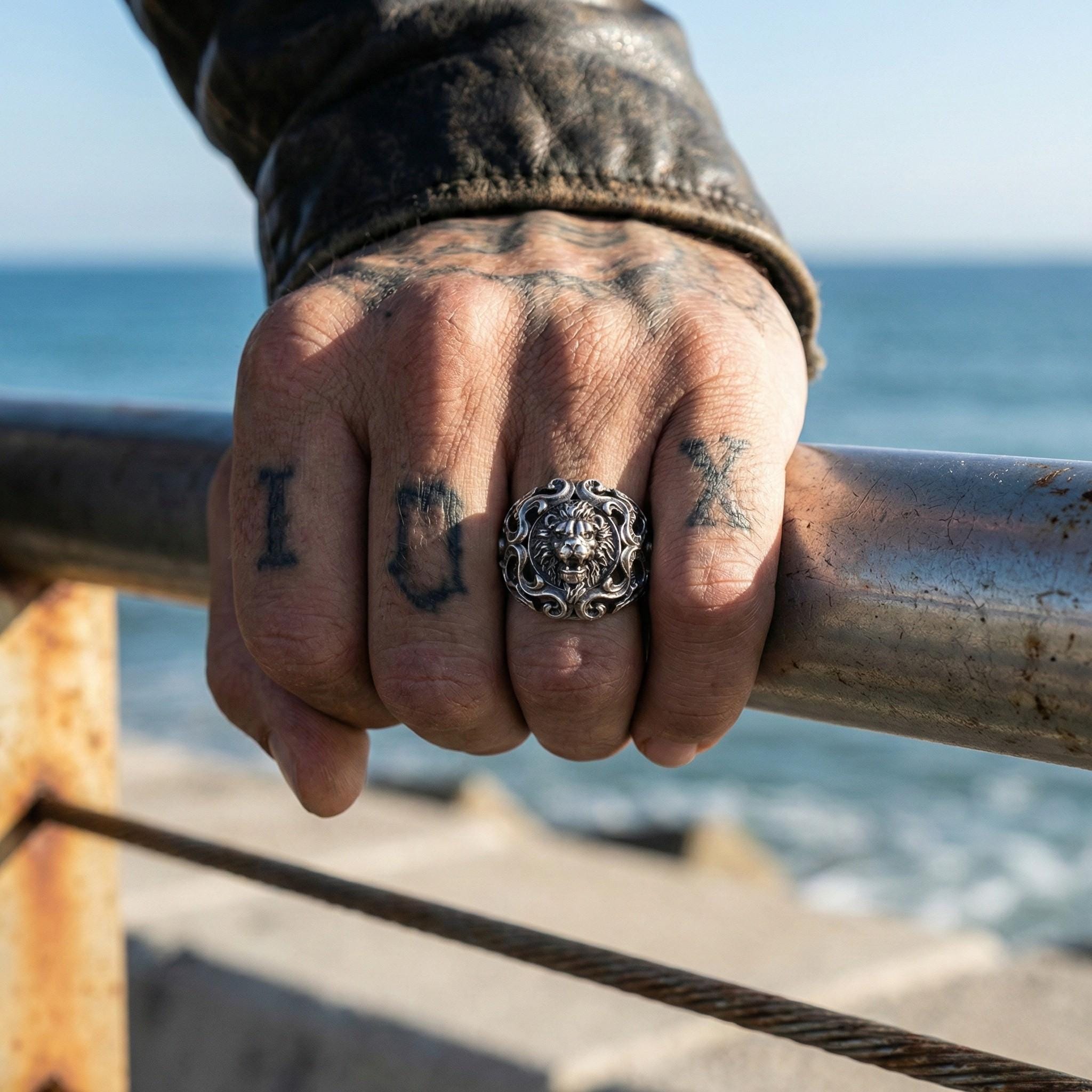 A close-up of a person's hand with tattoos on the fingers, wearing a ring, and holding onto a metal railing overlooking the ocean.