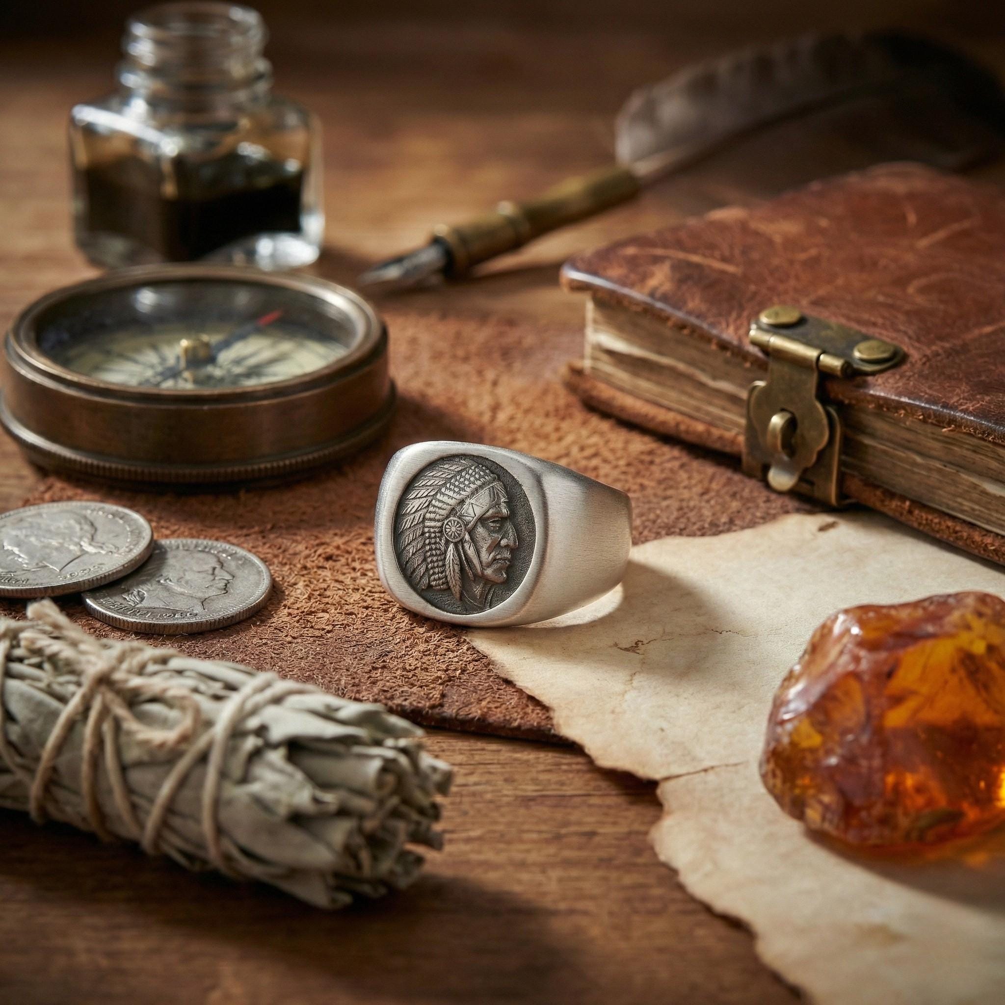a ring with a Native American head design, a compass, a book, a piece of paper, a coin, a piece of amber, and a small bottle of ink on a wooden surface.