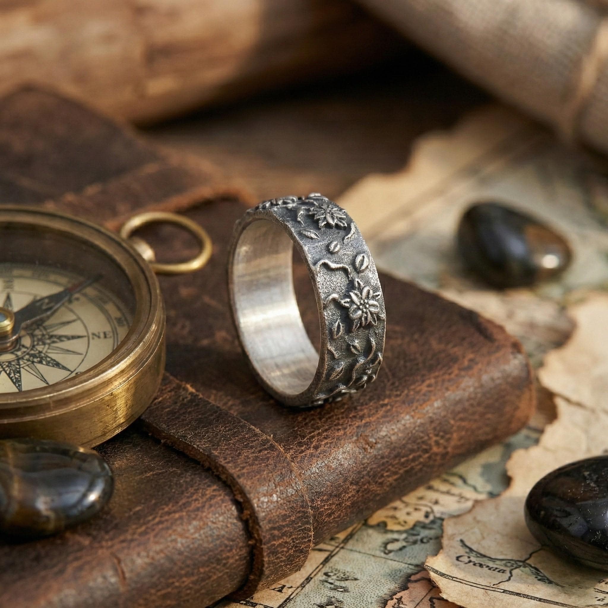 a silver ring with a floral design, placed on top of a leather-bound book or notebook, along with a compass and some stones.