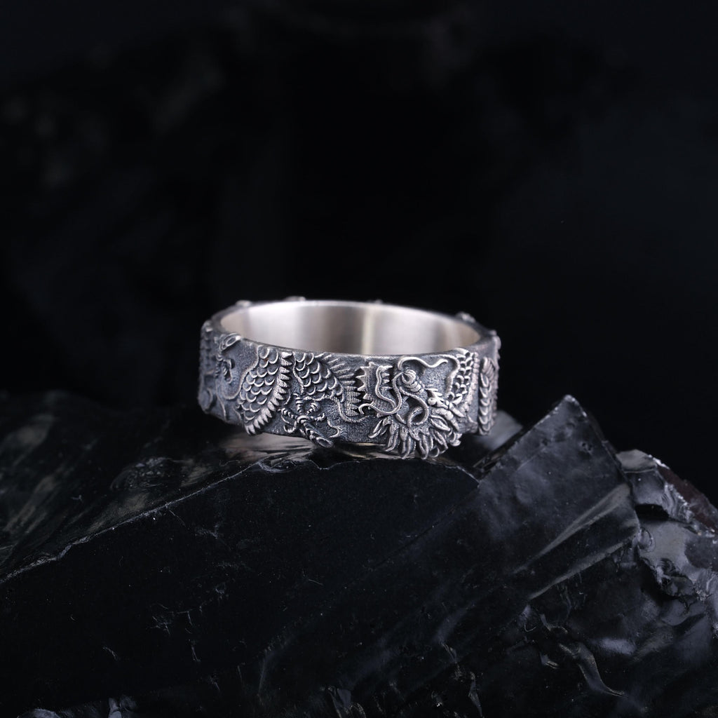 A silver ring with an intricate dragon design is displayed against a dark background.