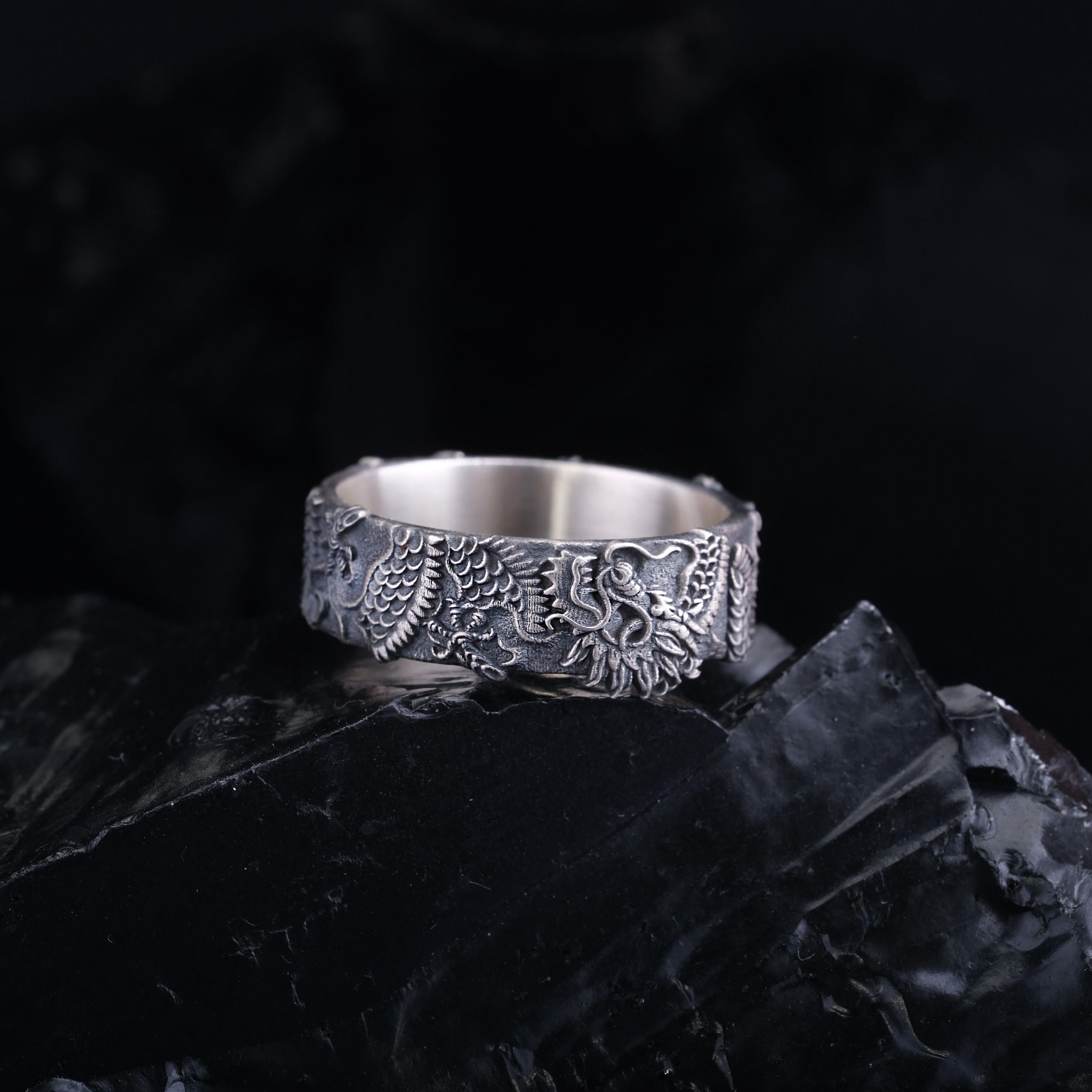A silver ring with an intricate dragon design is displayed against a dark background.