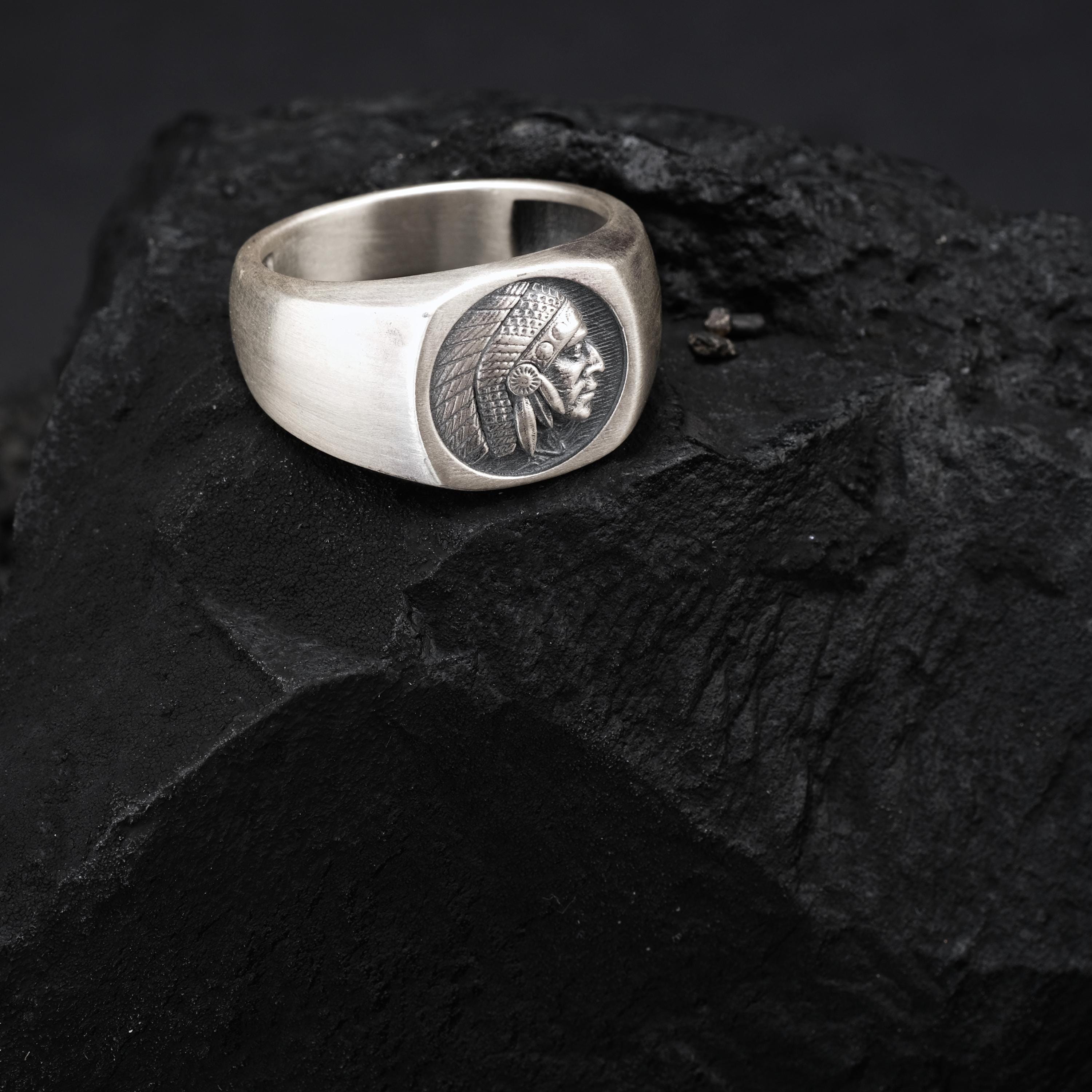 A silver ring with a carved design of a Native American head is resting on a black surface.