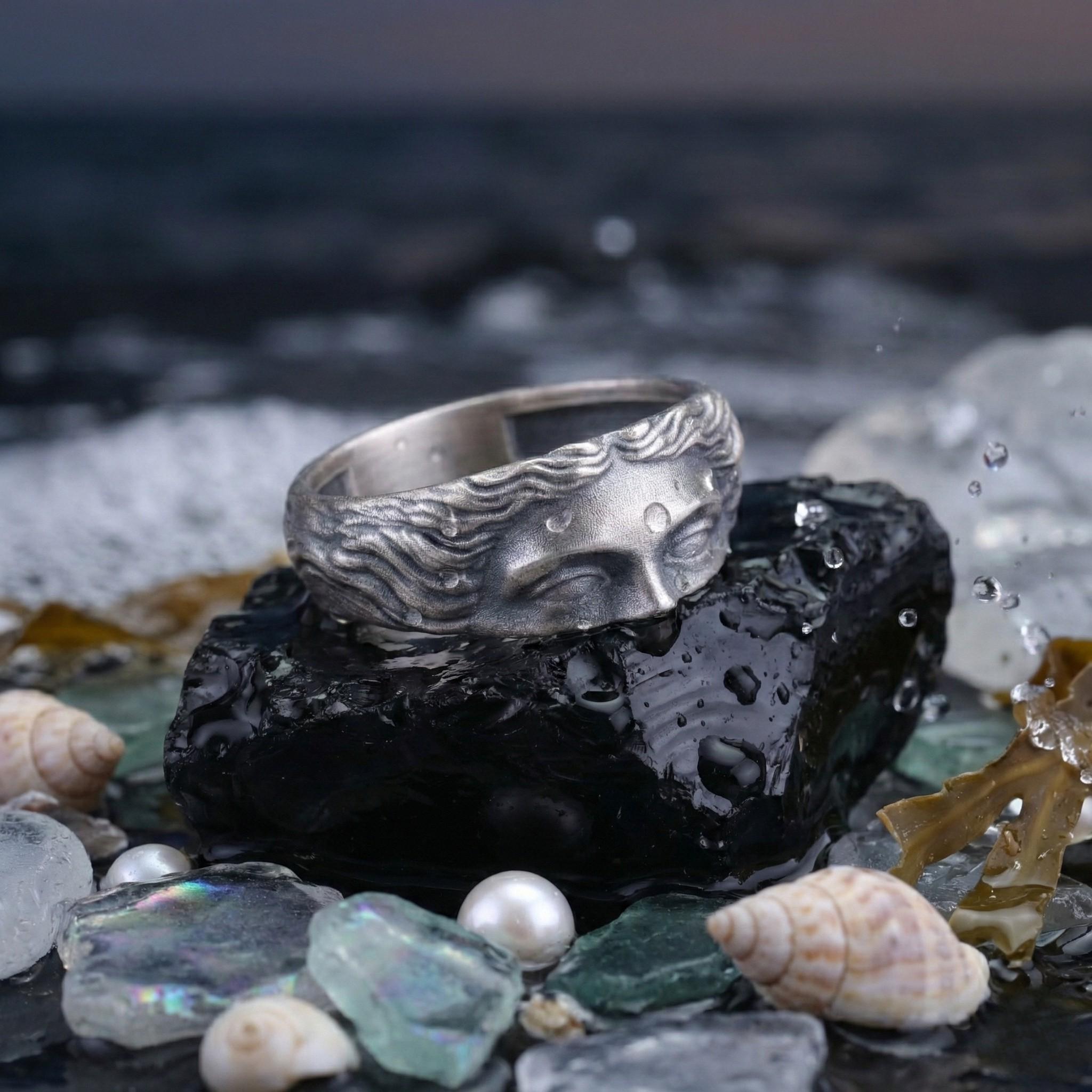 a silver ring with a face design, placed on a black surface surrounded by various sea-themed objects such as shells, pearls, and rocks.
