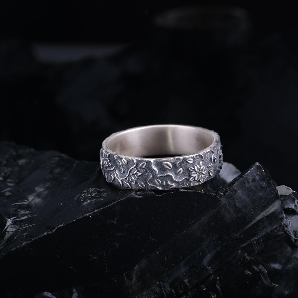 A silver ring with a floral design sits on a black surface against a dark background.