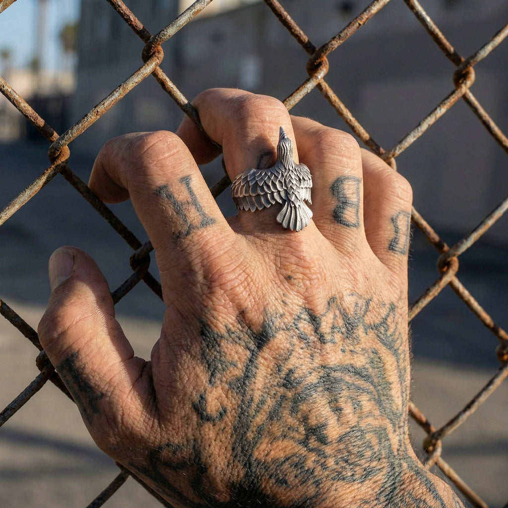 A close-up of a tattooed hand wearing a ring with a bird design, with a chain link fence in the background.