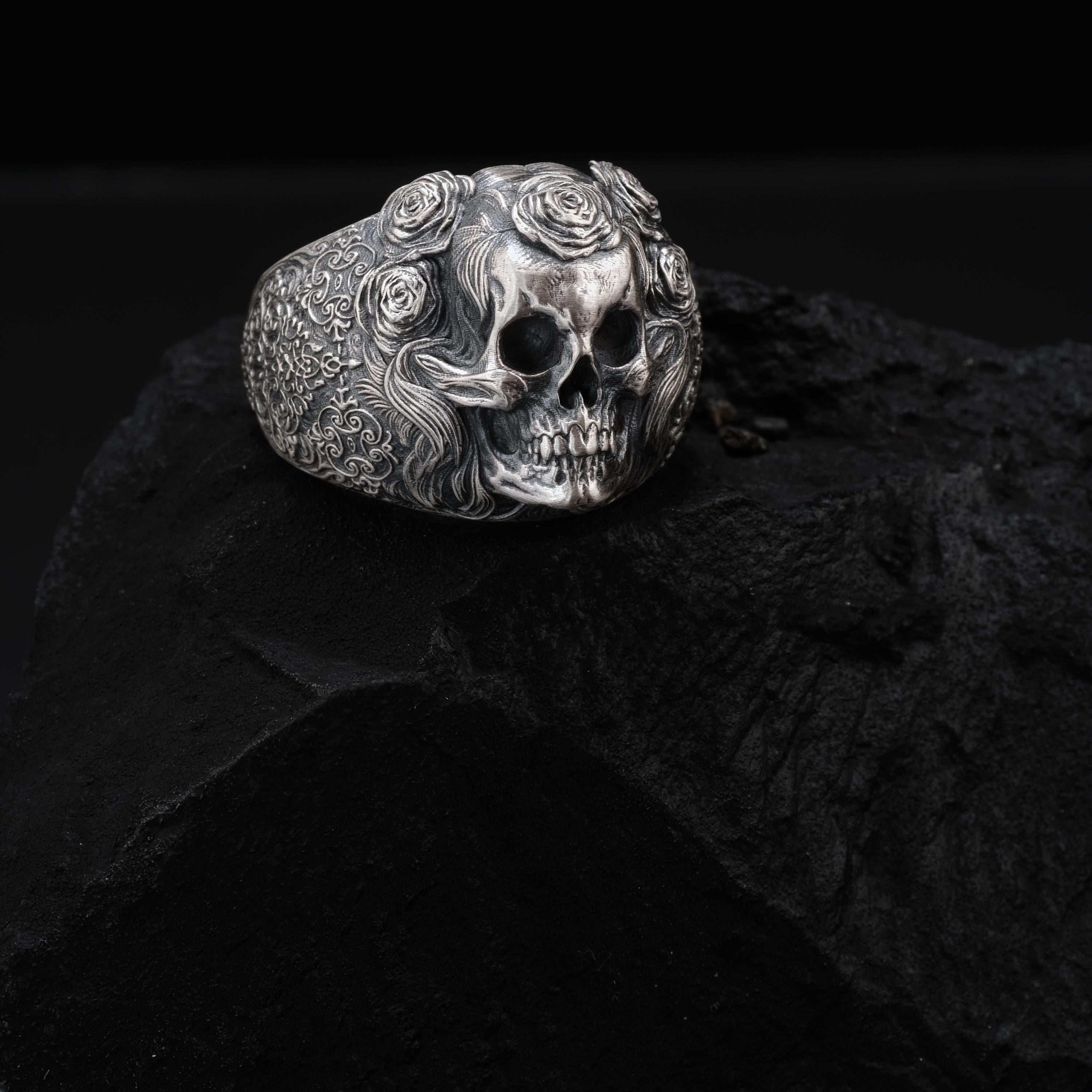 A silver ring with a skull design, featuring intricate floral patterns, is resting on a dark surface.