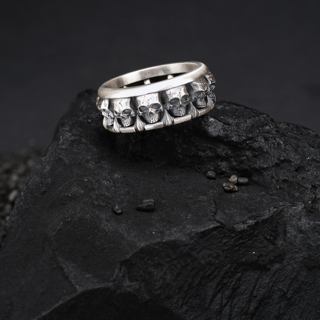 A silver ring with multiple skull designs on it, resting on a dark surface.