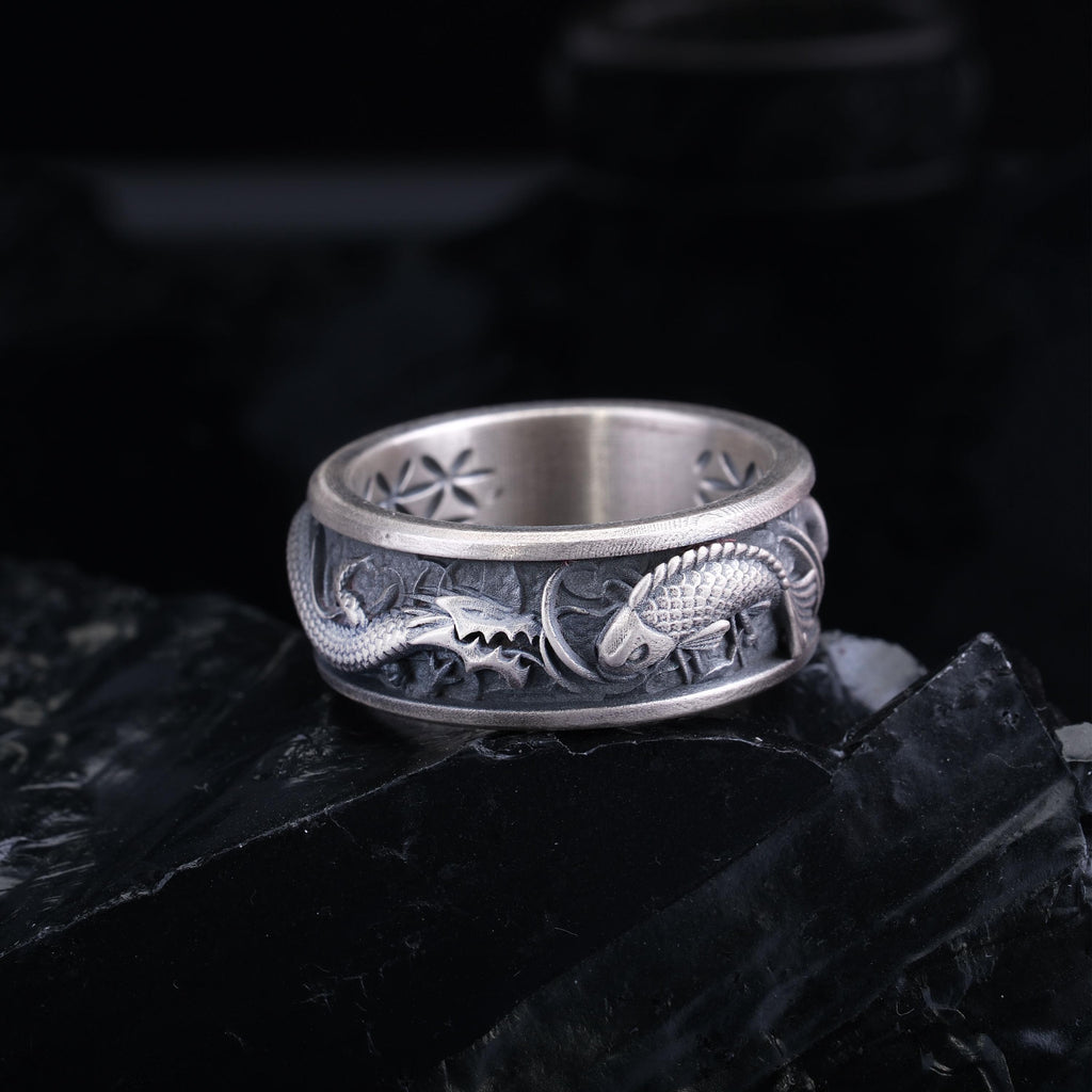 A silver ring with an intricate dragon design on the band, resting on a black surface.