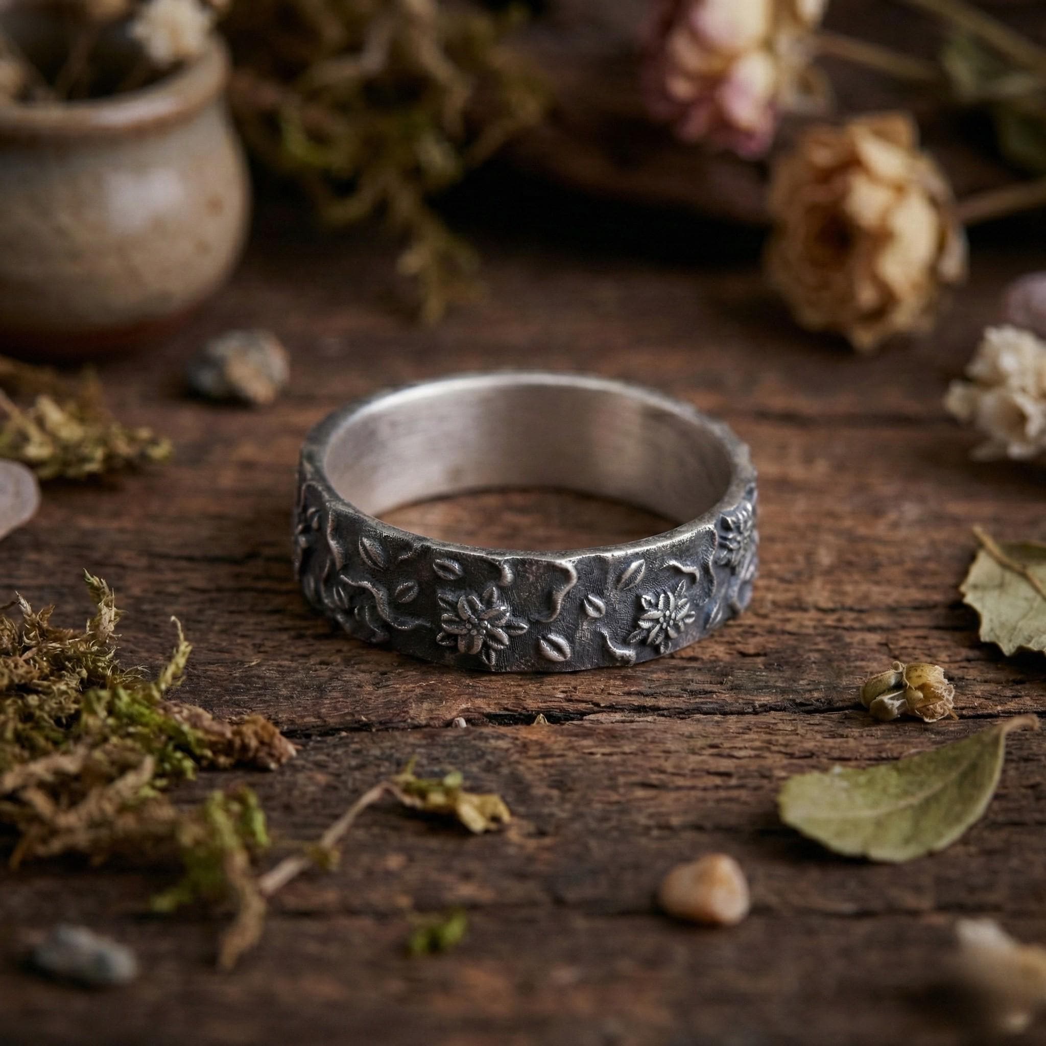 A silver ring with a floral design sits on a wooden surface, surrounded by dried flowers, leaves, and other natural elements.