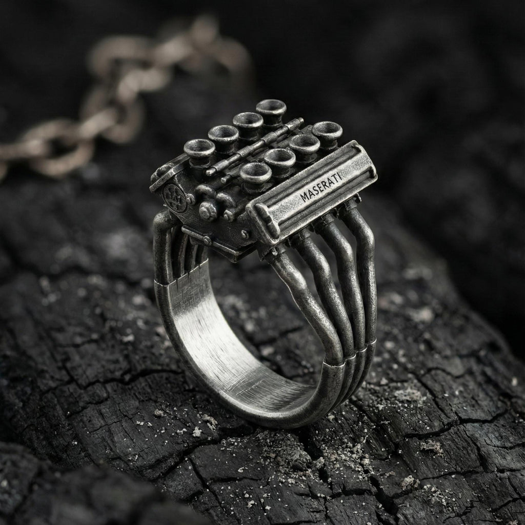 A silver ring with a unique design, featuring a metallic structure resembling an engine or a car part, placed on a dark, textured surface.