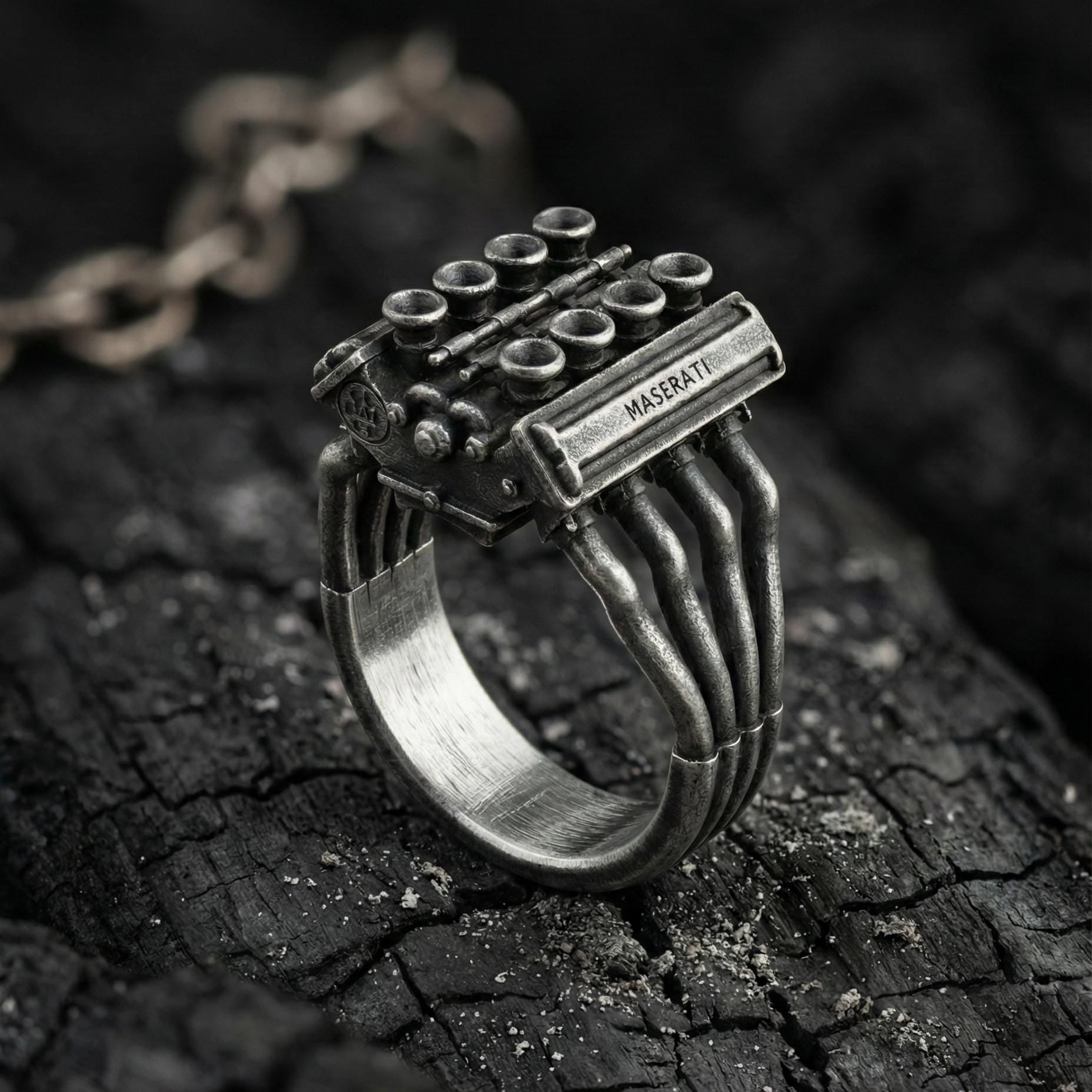A silver ring with a unique design, featuring a metallic structure resembling an engine or a car part, placed on a dark, textured surface.