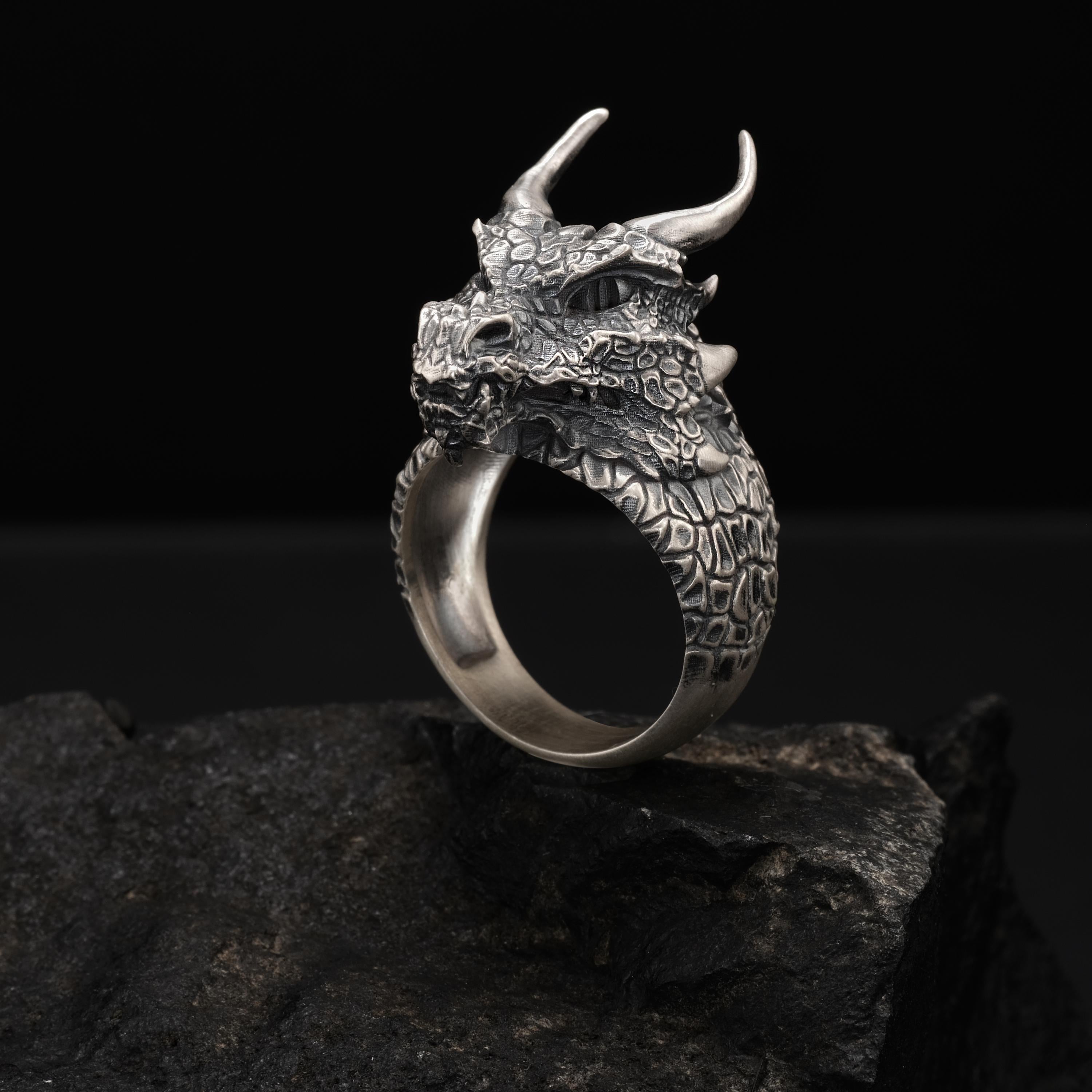 A silver ring with a dragon head design is displayed on a rock against a dark background.