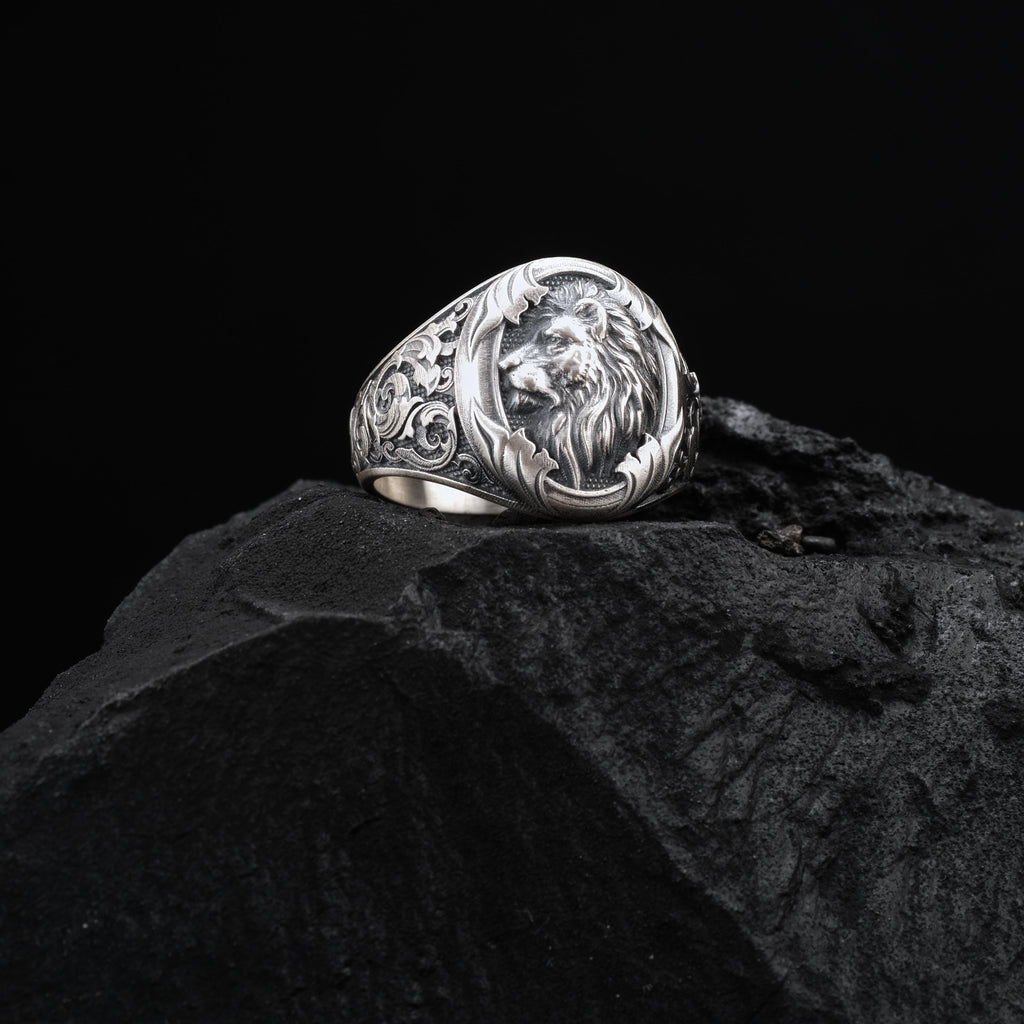 A silver ring with an intricate lion head design sits atop a dark, rocky surface.