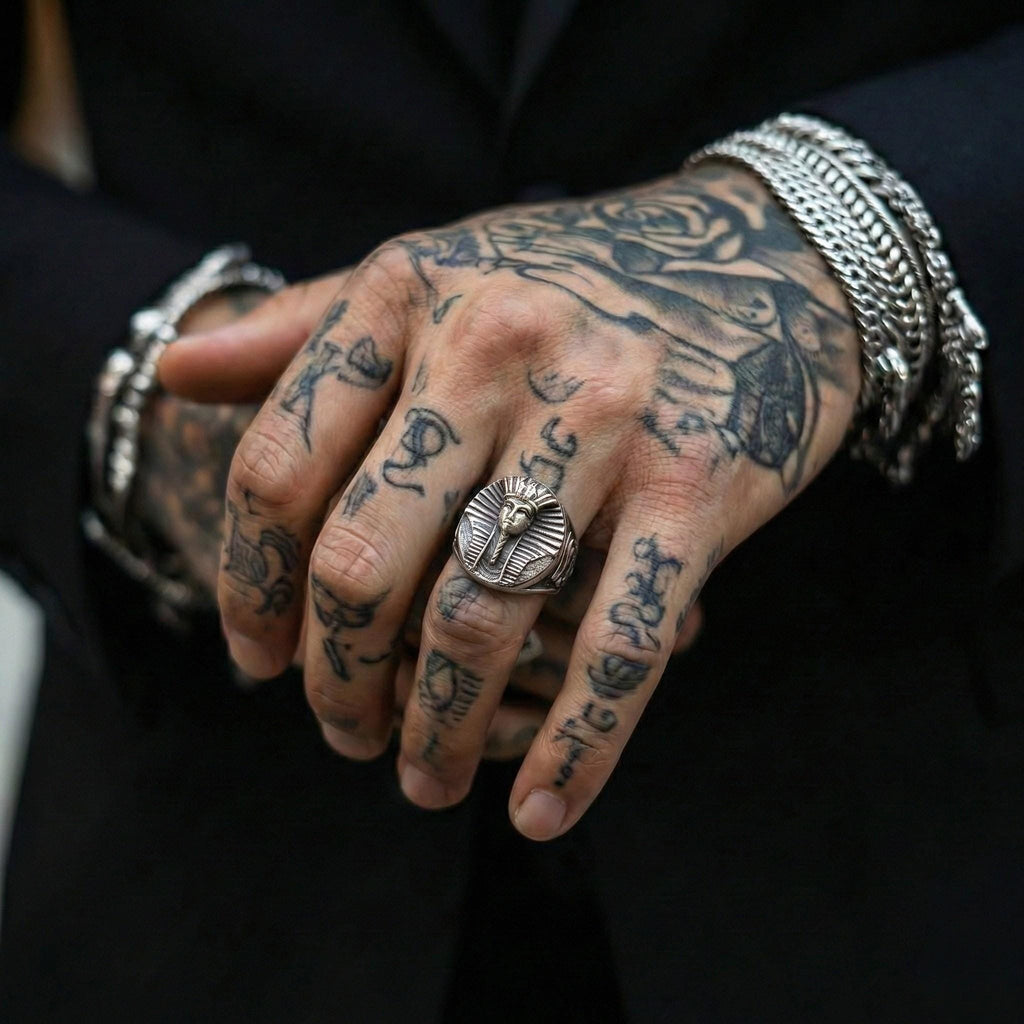 A close-up of a person's hand with a large, ornate ring on the finger, surrounded by various tattoos on the fingers and palm area.