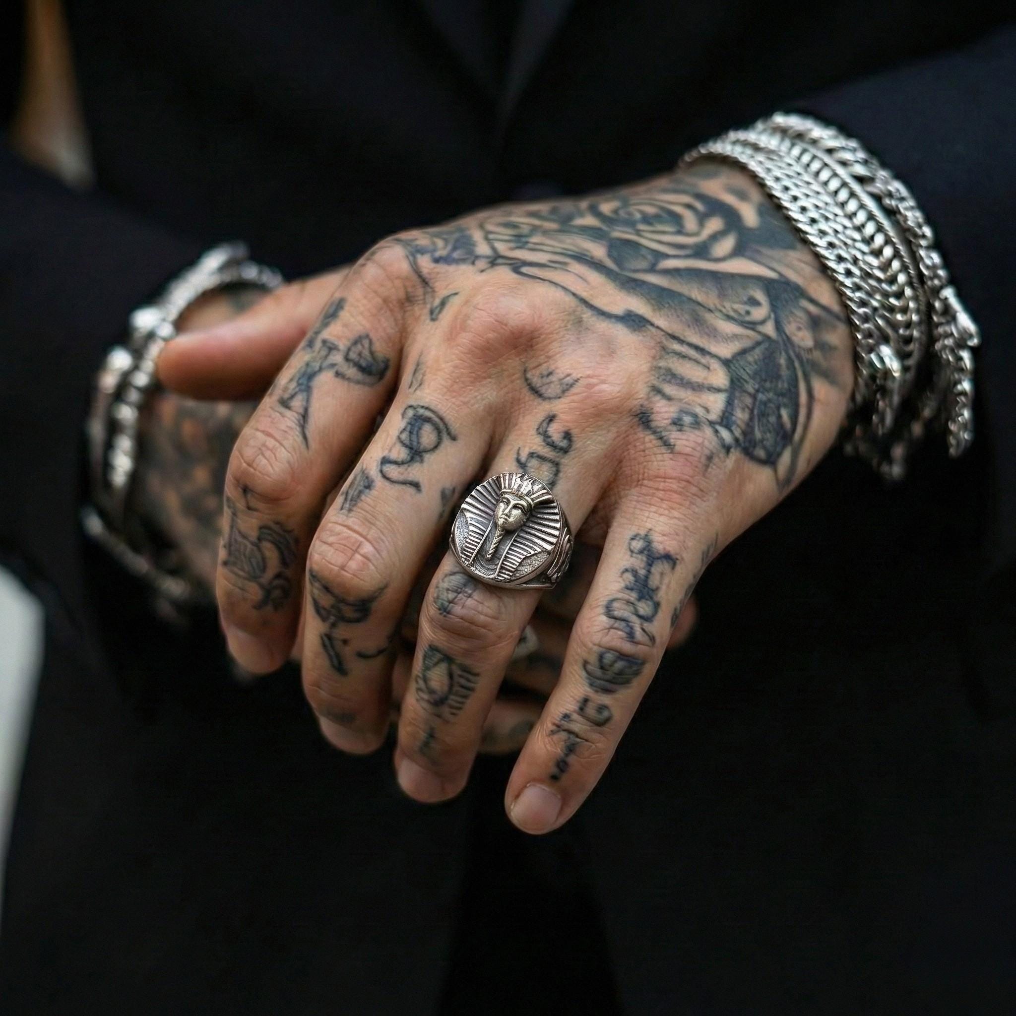A close-up of a person's hand with a large, ornate ring on the finger, surrounded by various tattoos on the fingers and palm area.