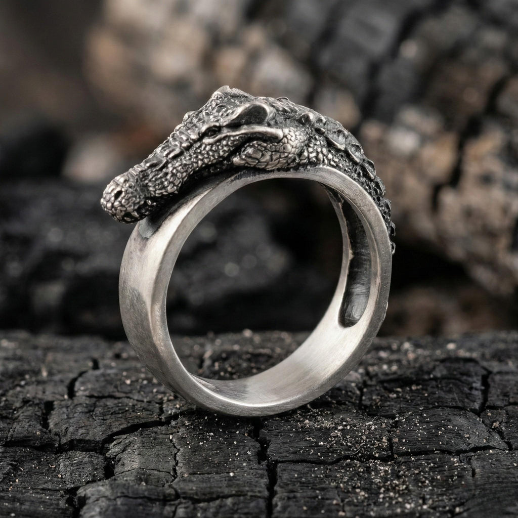 A silver ring with a detailed crocodile head design is displayed on a dark, cracked surface.