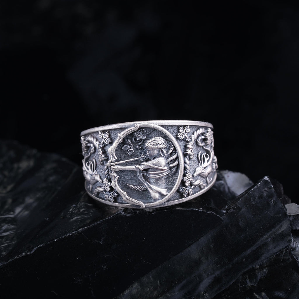 A silver ring with an intricate design featuring a figure holding a bow and arrow, surrounded by floral and geometric patterns.