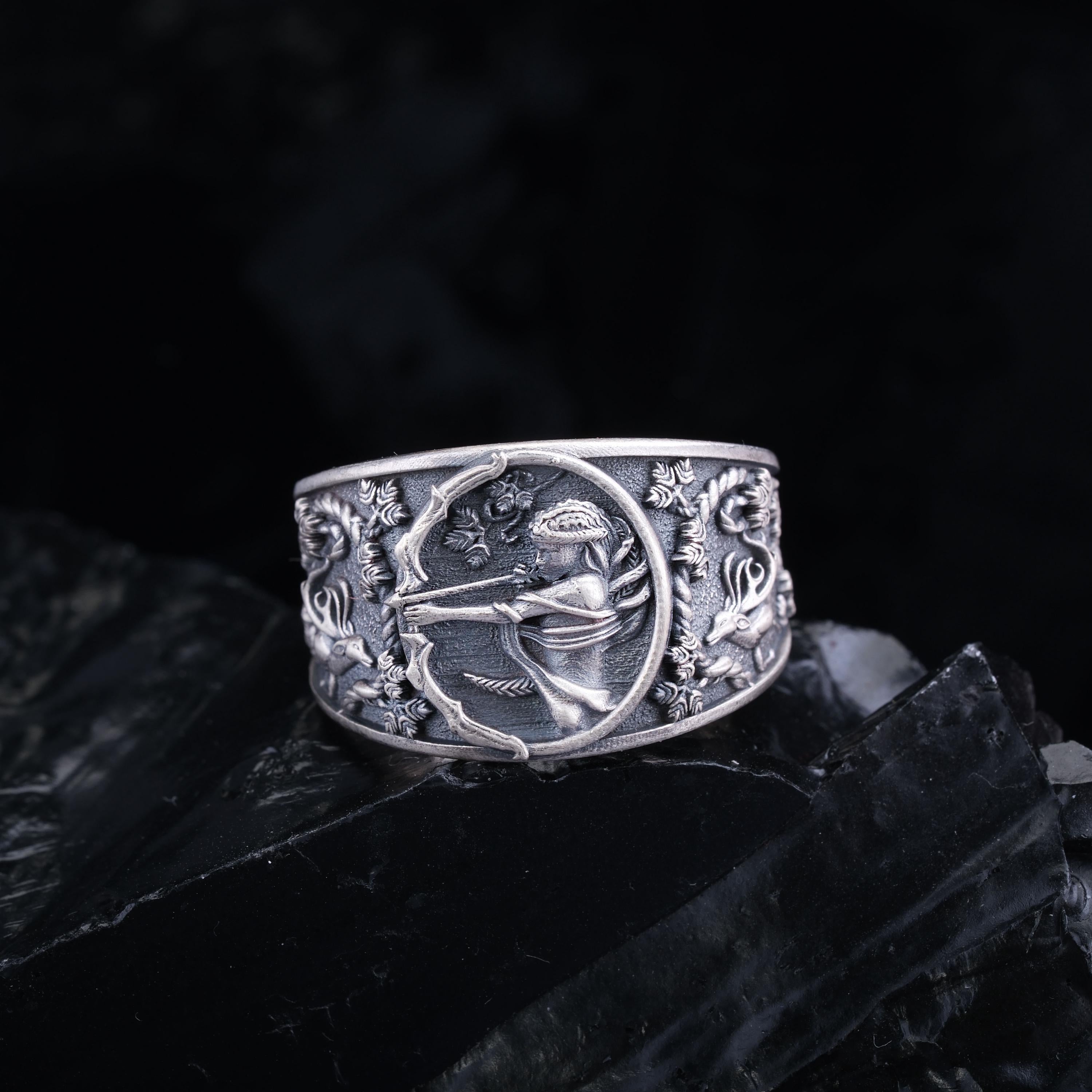 A silver ring with an intricate design featuring a figure holding a bow and arrow, surrounded by floral and geometric patterns.