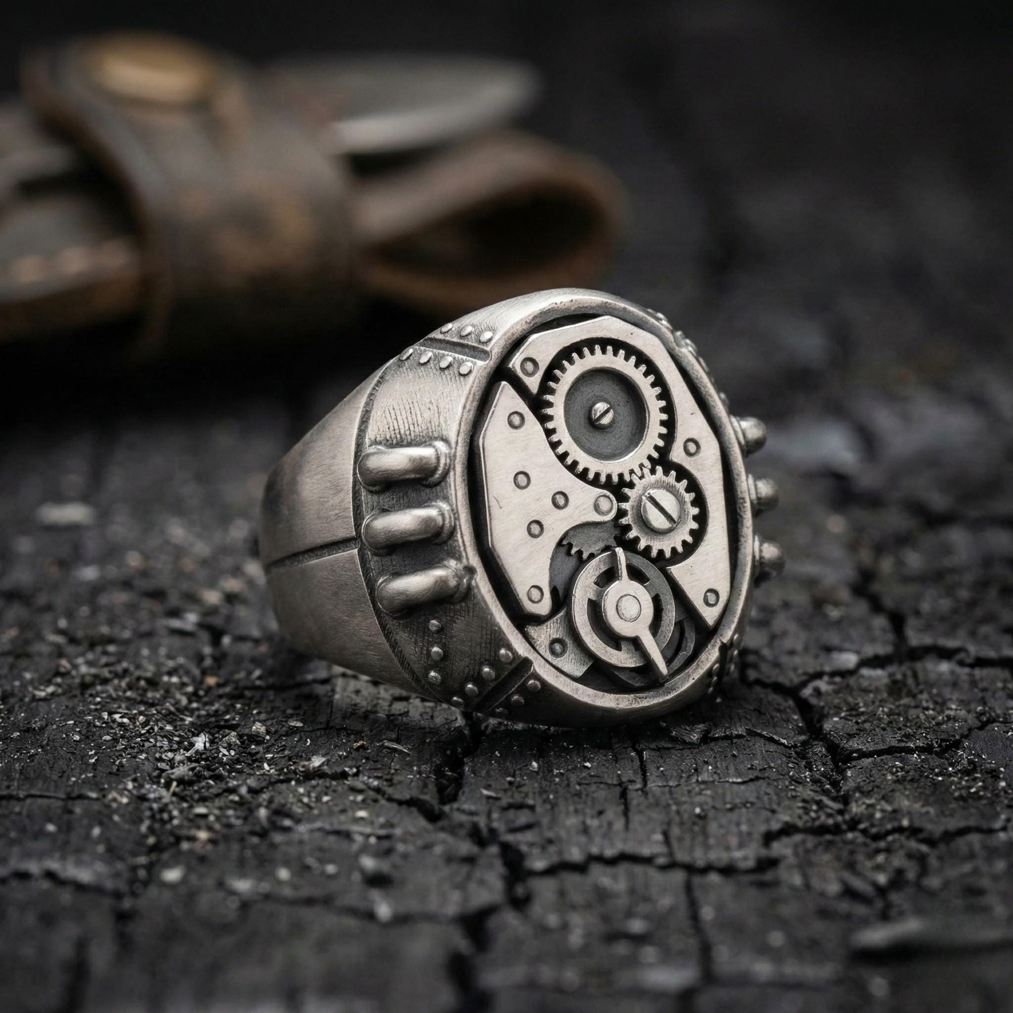 A close-up image of a silver ring with a mechanical or industrial design, resting on a dark, cracked surface.