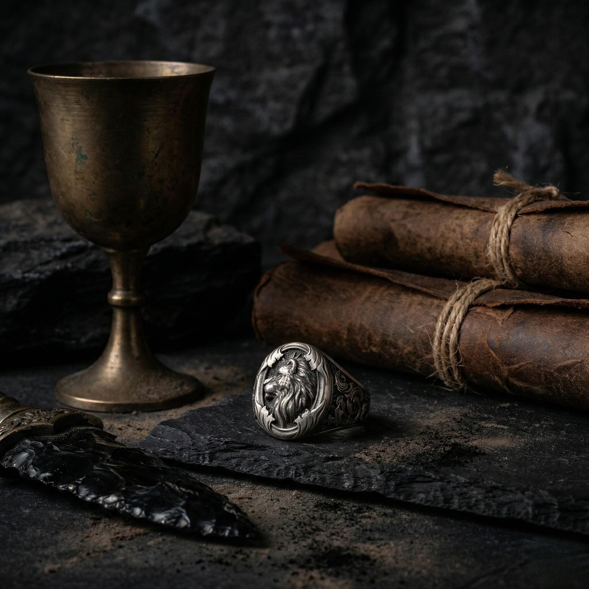 a ring with a lion design, a metal cup, a scroll, a book, and a stone or rock, all arranged on a dark surface.