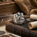 a silver ring with a shield design, placed on top of a leather-bound book, surrounded by coins and a feather.