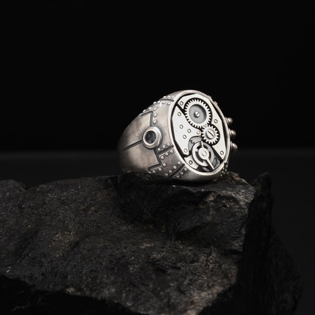 A silver ring with intricate mechanical designs sits on a dark rock.