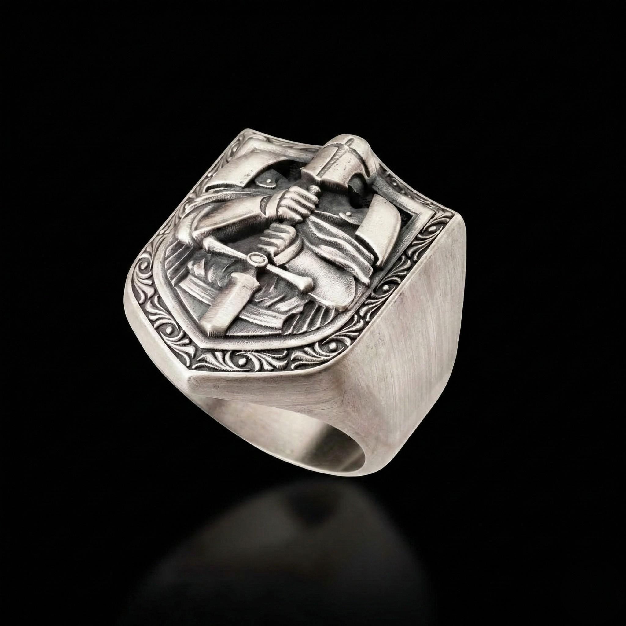 A silver ring with an intricate design featuring a shield-shaped motif, possibly depicting a knight or warrior figure, and a sword.