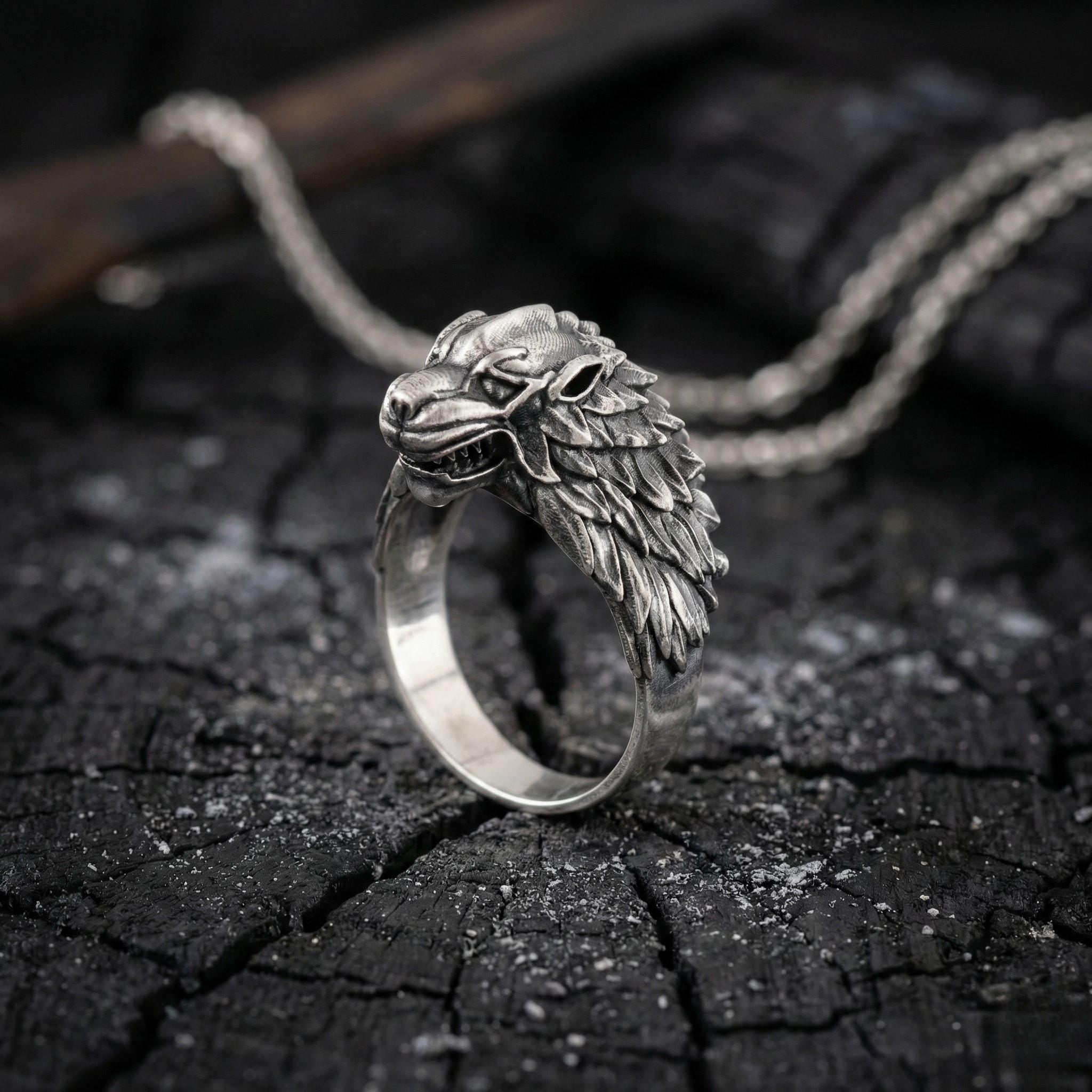 A silver ring with a detailed wolf head design is displayed on a dark, cracked surface.