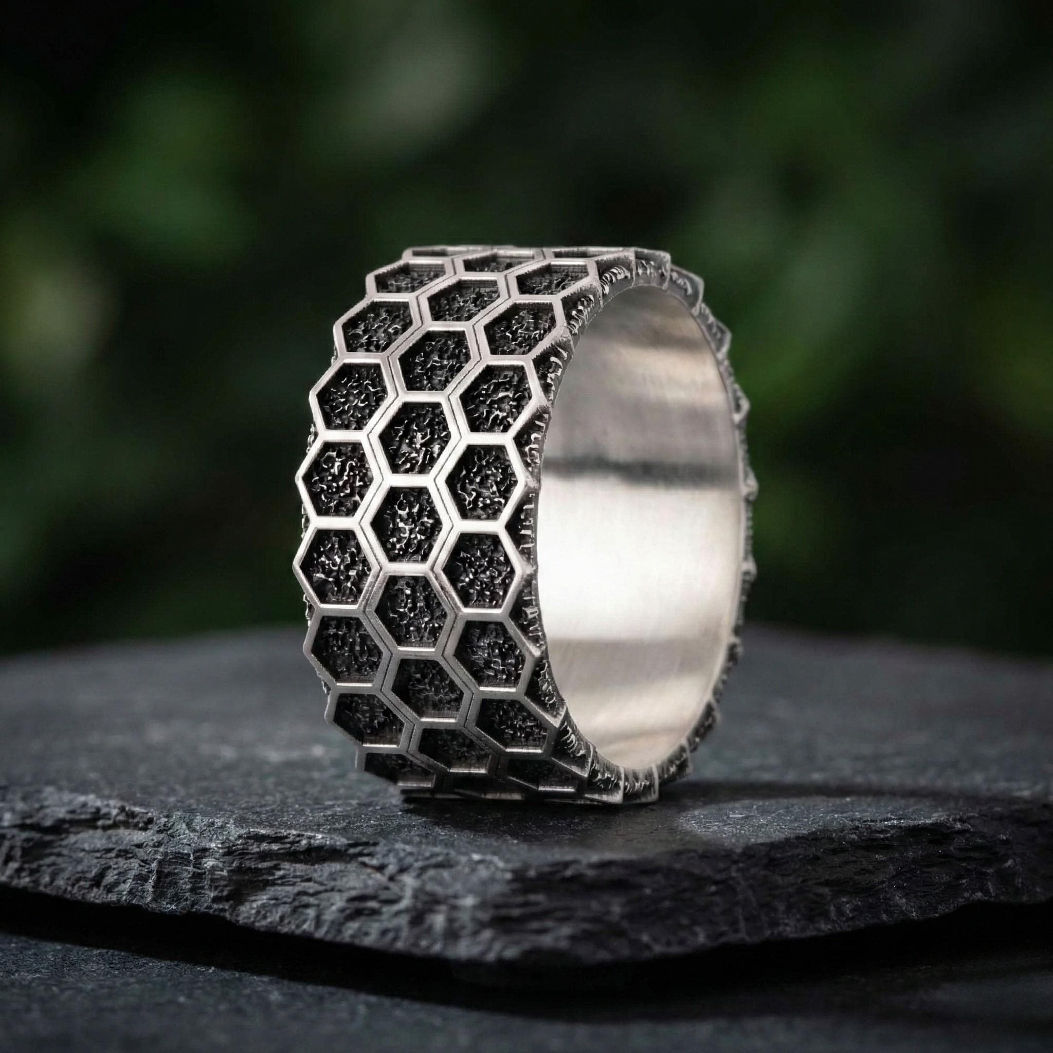 A silver ring with a honeycomb pattern design, featuring small black hexagonal shapes, is displayed on a dark surface.