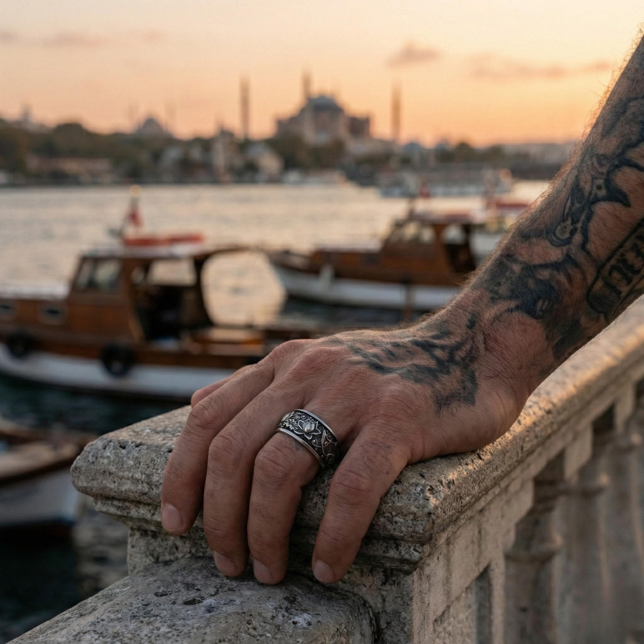 A hand with a tattoo on the arm is resting on a stone railing, with a view of a city skyline and boats in the background.