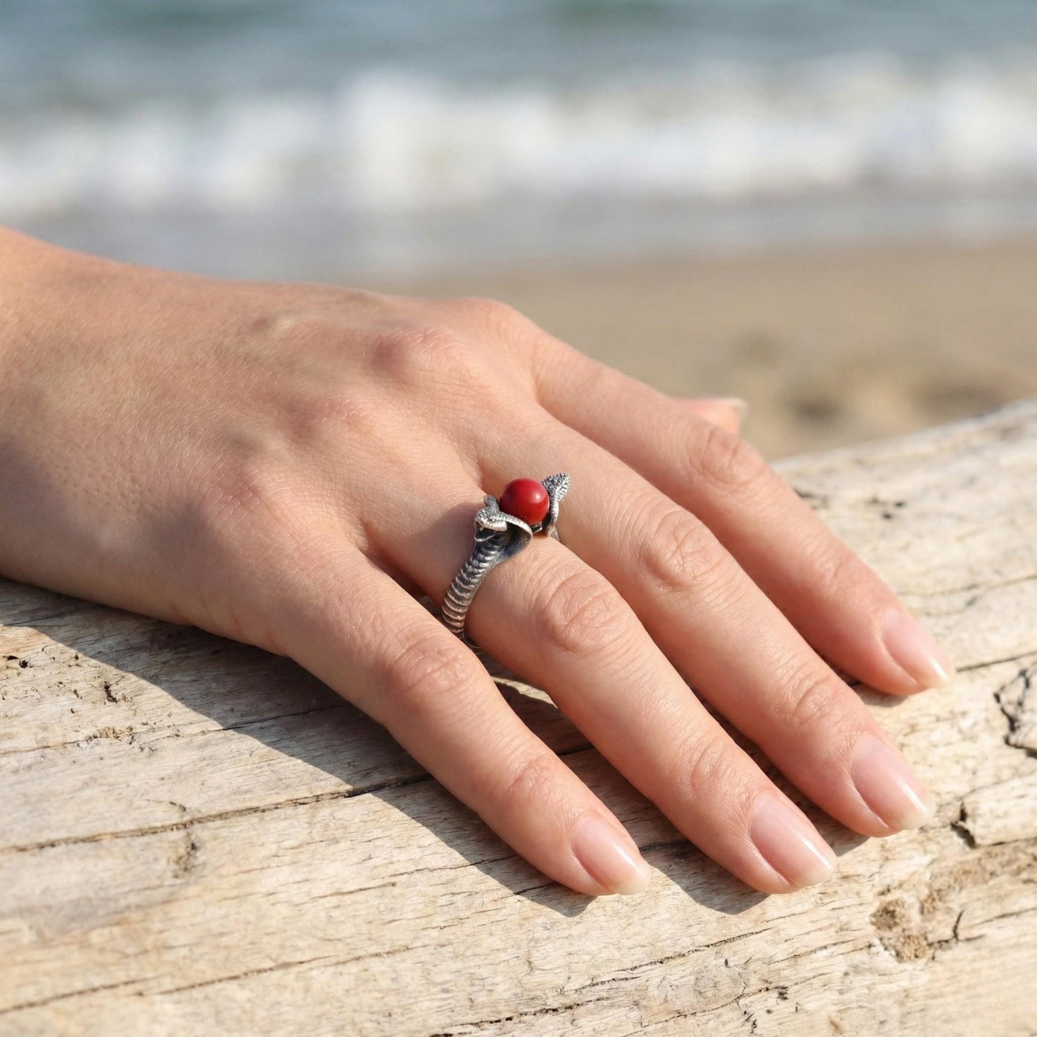 A close-up of a person's hand wearing a ring with a red gemstone, resting on a wooden surface with a blurred background of the ocean.