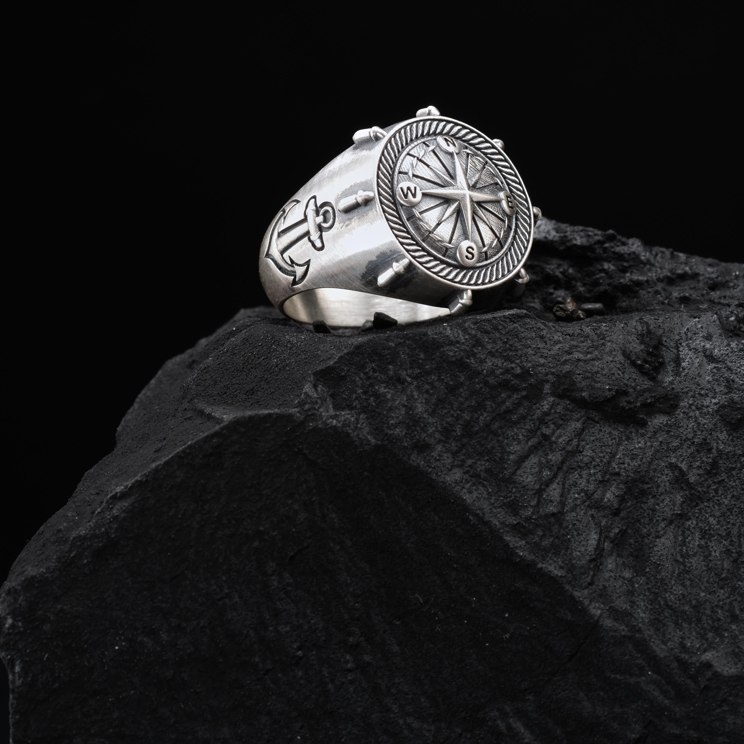 A silver ring with an anchor and compass design sits on a black rock.
