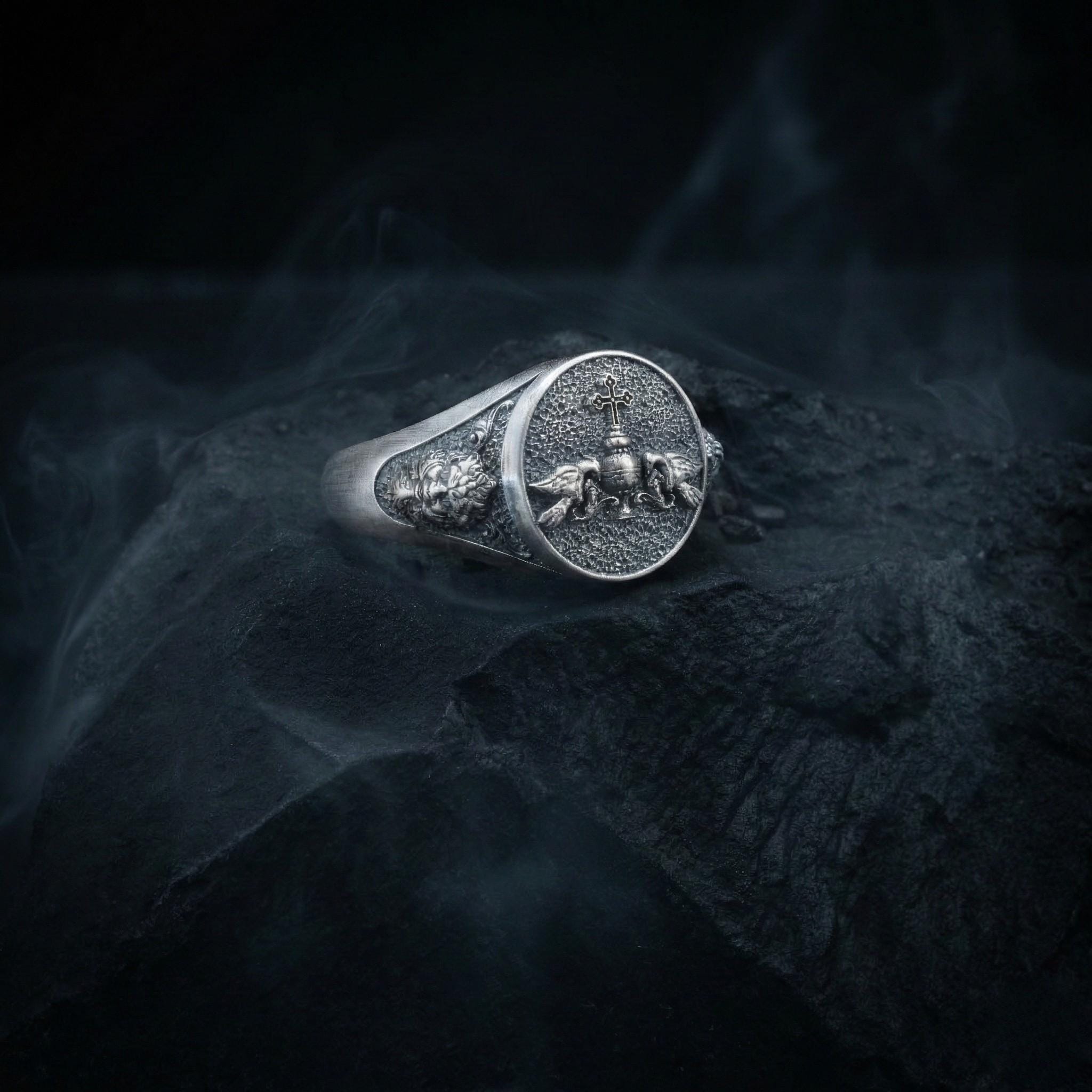 A silver ring with a detailed design sits on a dark surface, surrounded by smoke.