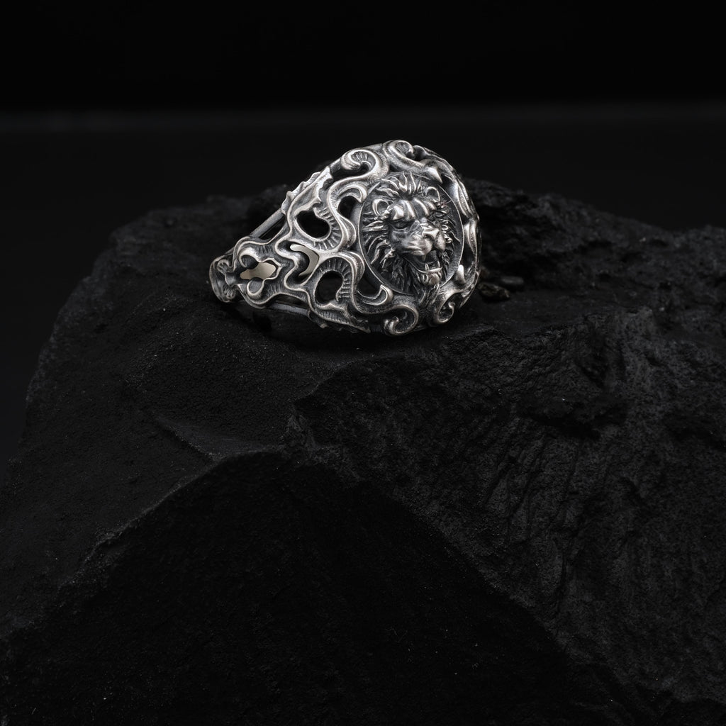 A silver ring with an intricate design sits on a dark surface, possibly a rock or a piece of charcoal.